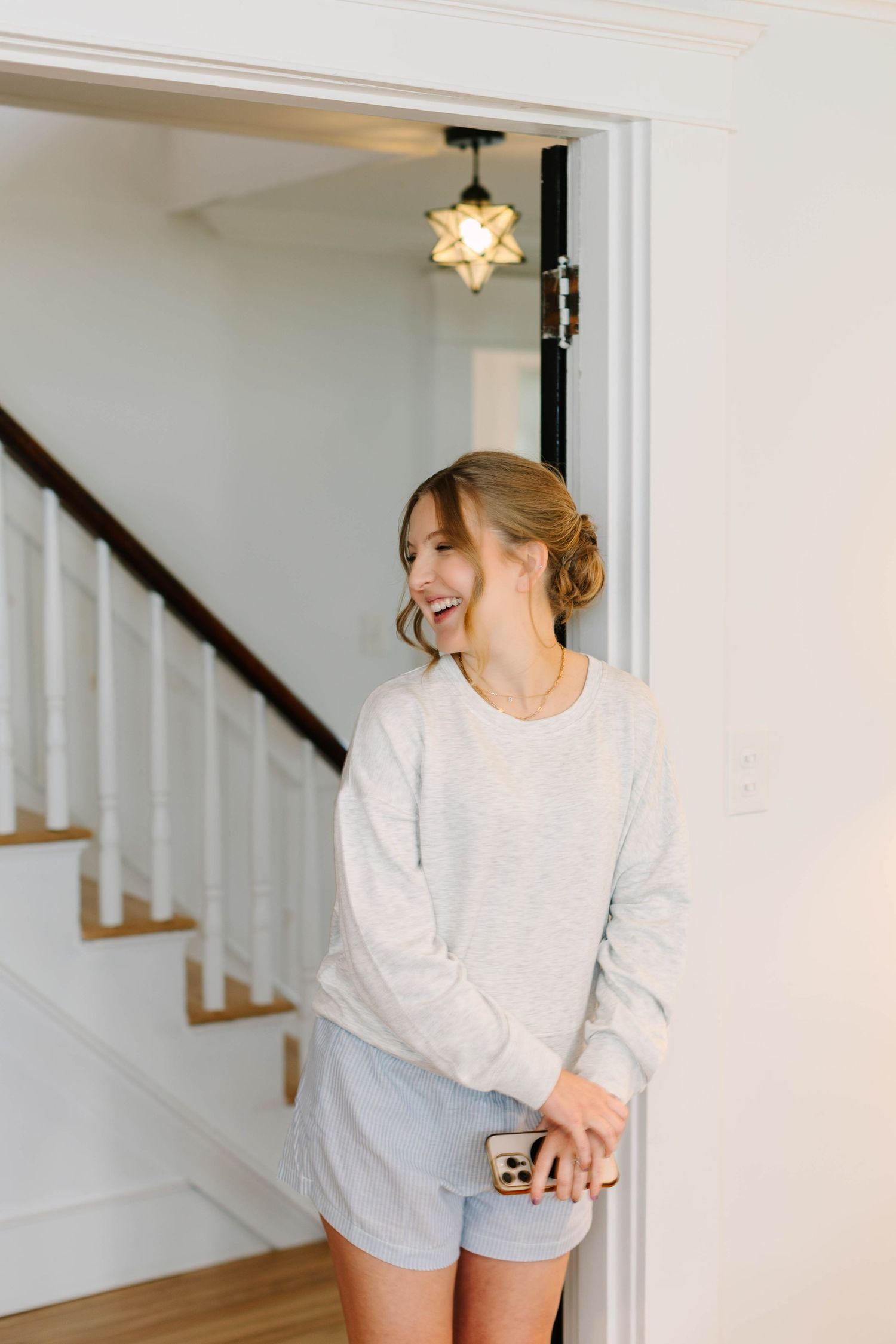 A smiling woman in a light gray sweatshirt and shorts holds a phone while standing in a bright white doorway near a staircase.