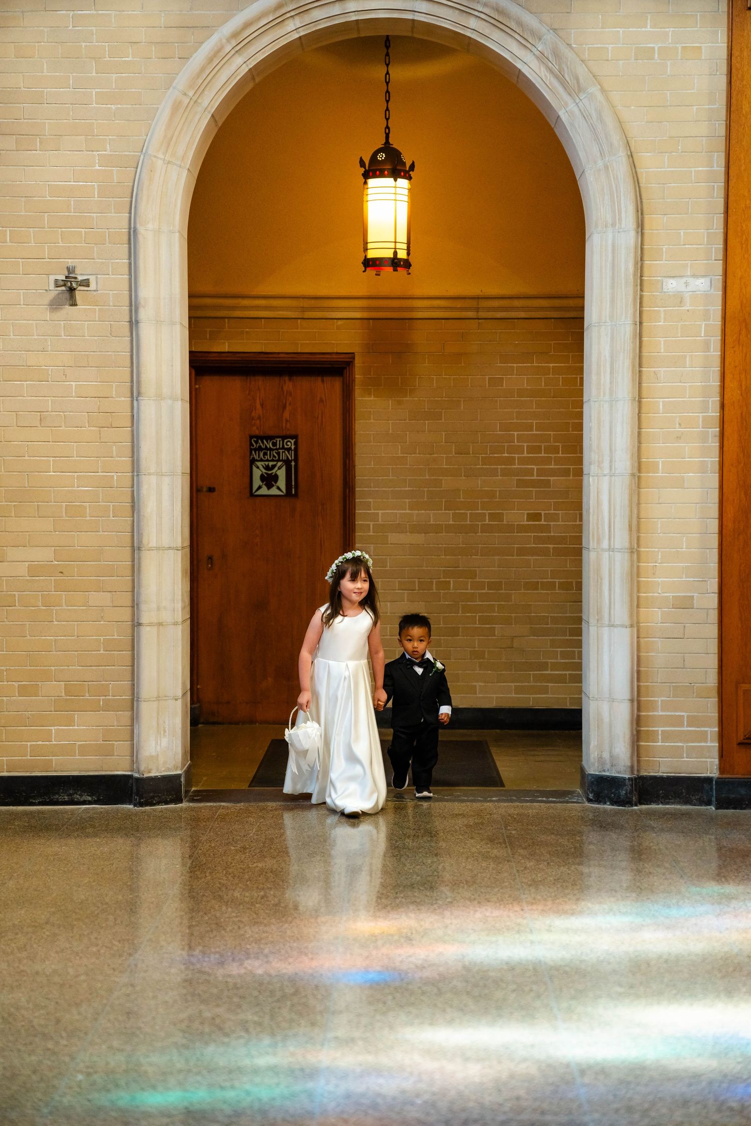 A flower girl in white dress and ring bearer in tuxedo walk through an arched church entrance during a wedding ceremony.
