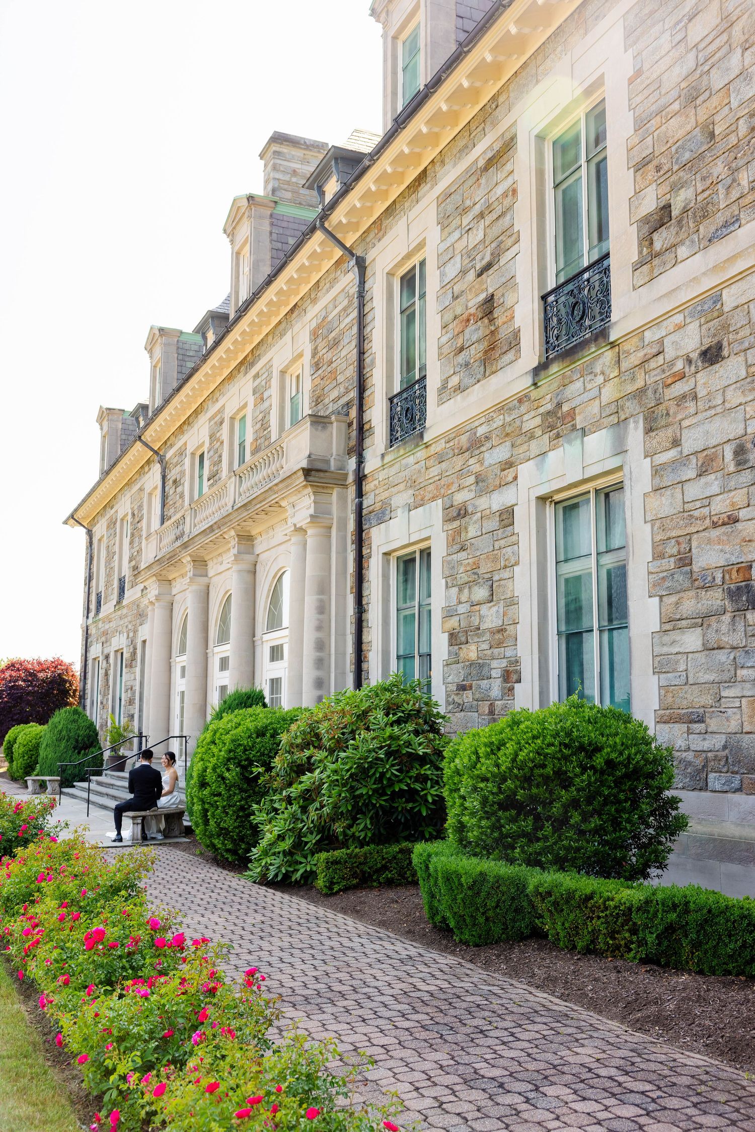 Grand stone mansion exterior with manicured hedges and colorful flower garden along cobblestone path.