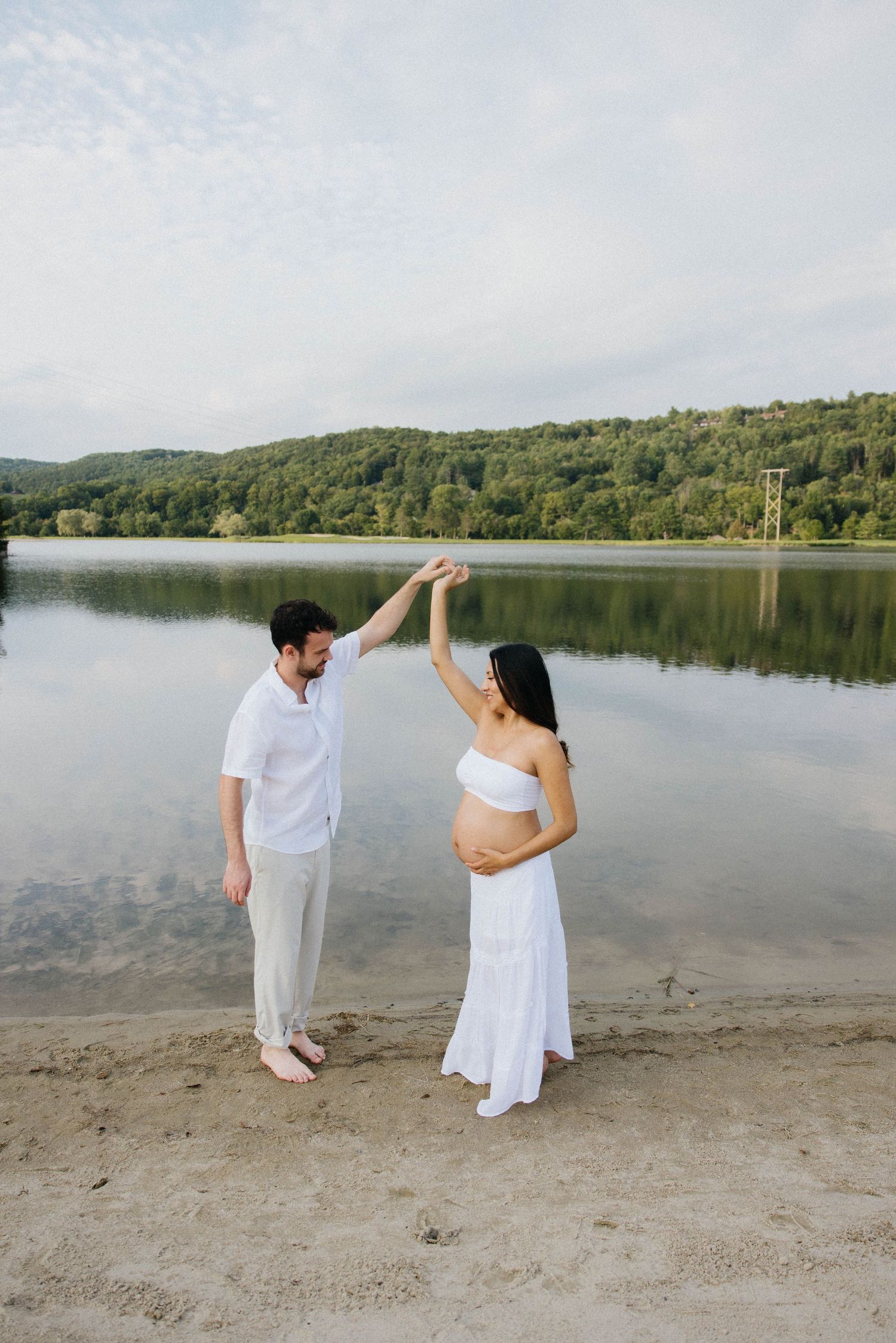 Couple in white outfits dancing on a sandy lakeside beach, pregnant woman in white dress, lush green hills in background.