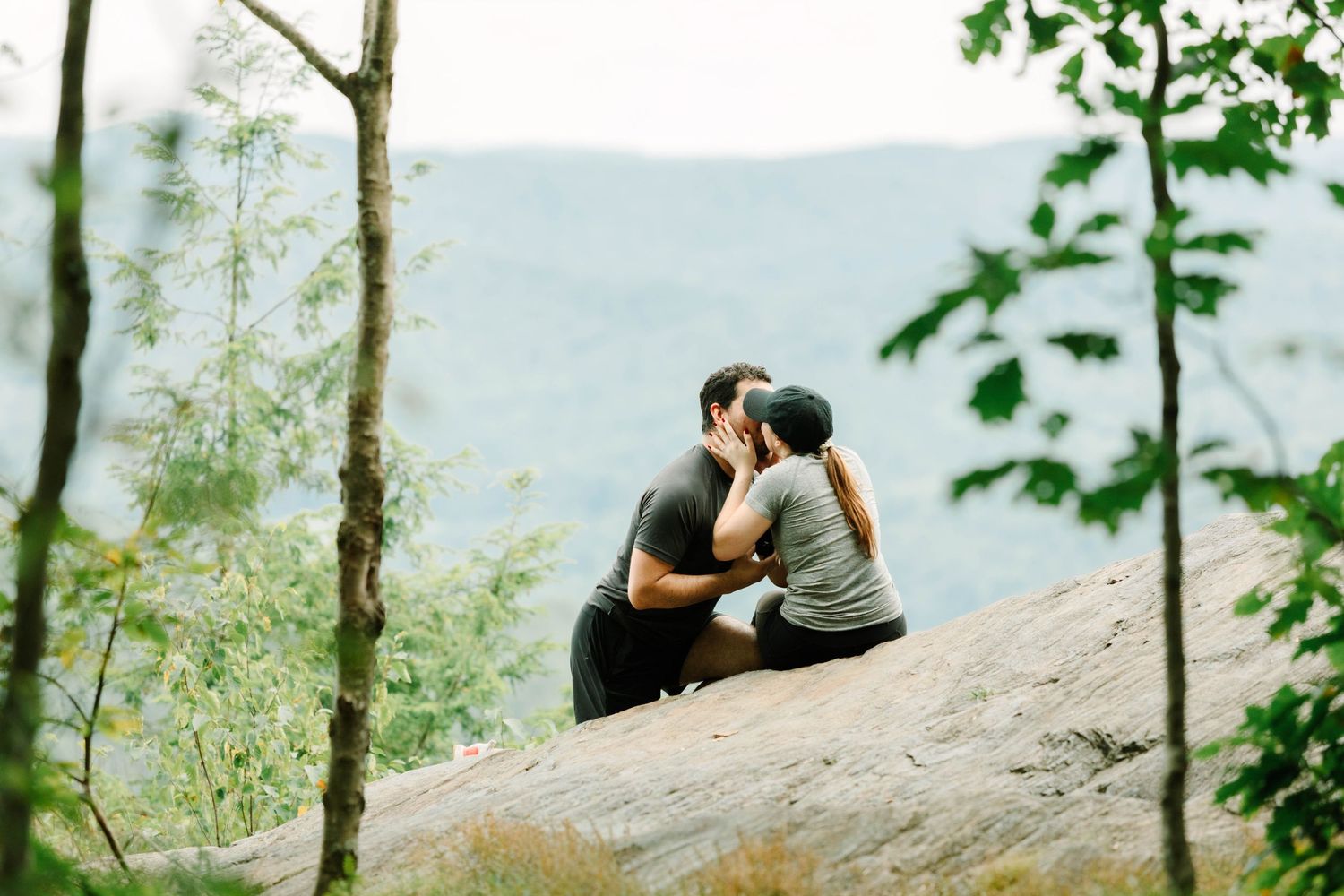 Couple sharing a romantic kiss on a rocky overlook surrounded by lush green trees and misty mountain views.