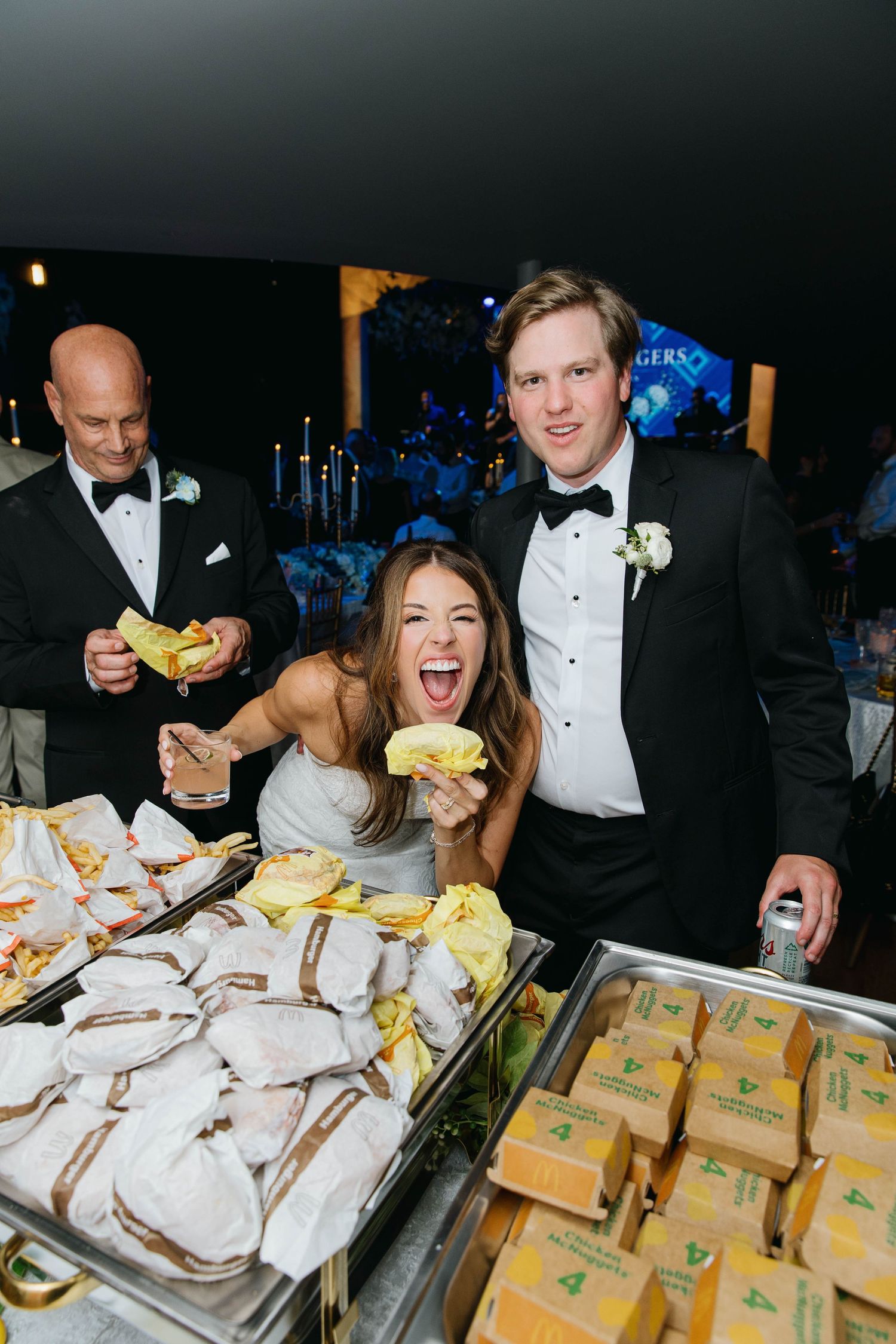 Bride and groom laughing at a wedding reception late-night snack buffet with wrapped sandwiches and cheeseburgers.