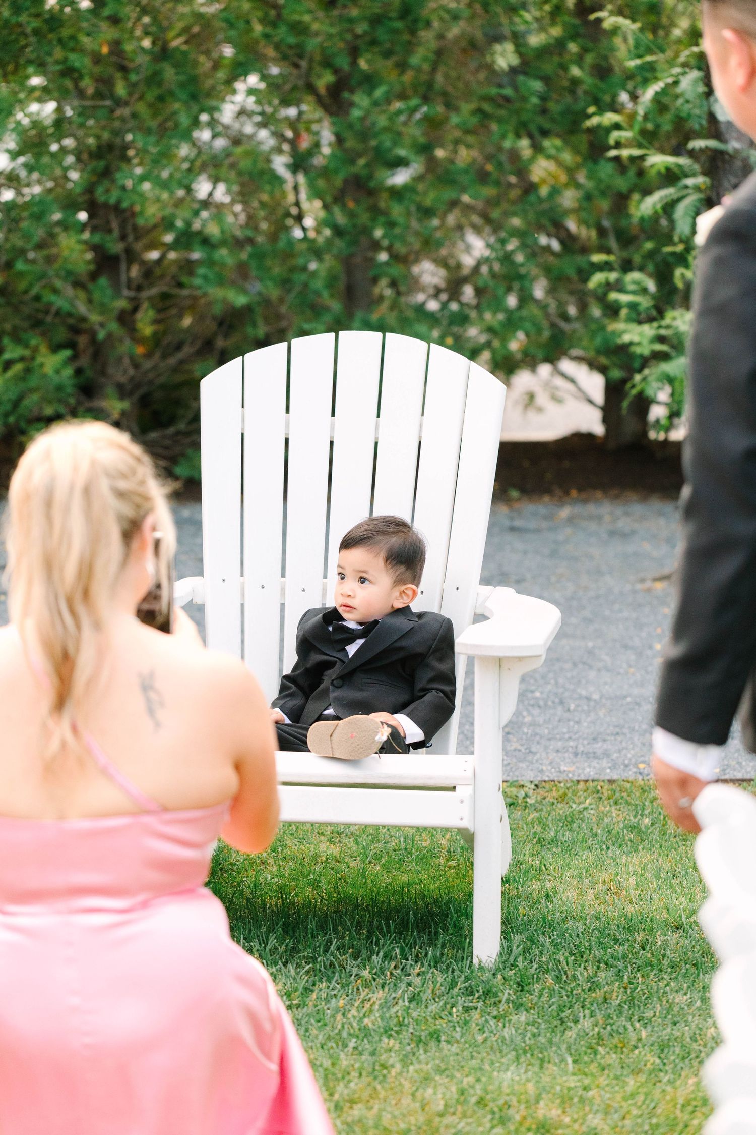 Adorable baby in black tuxedo sits in white Adirondack chair at outdoor wedding ceremony.
