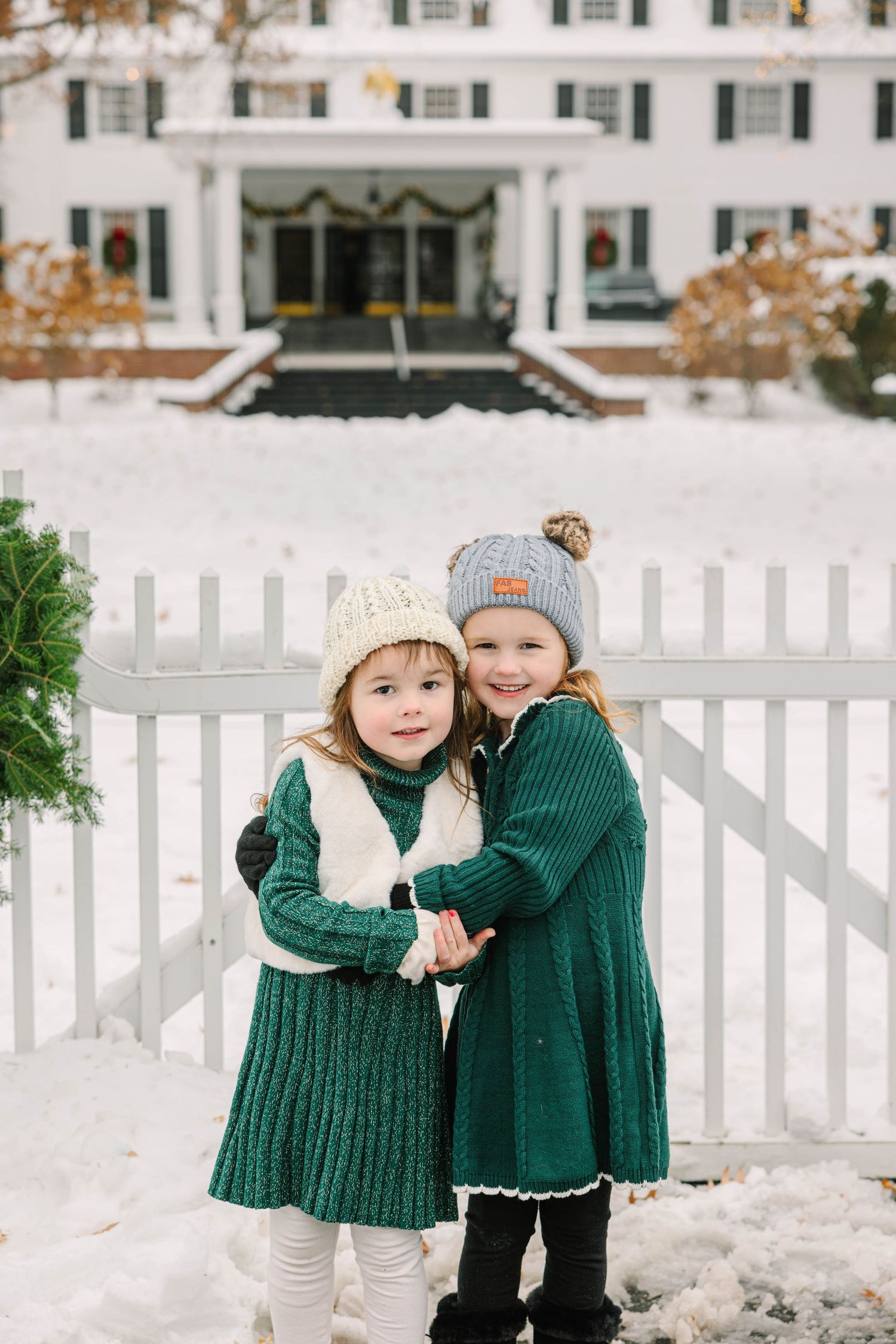 Two young girls in matching green knit dresses and winter hats embrace in front of a snow-covered white colonial building.