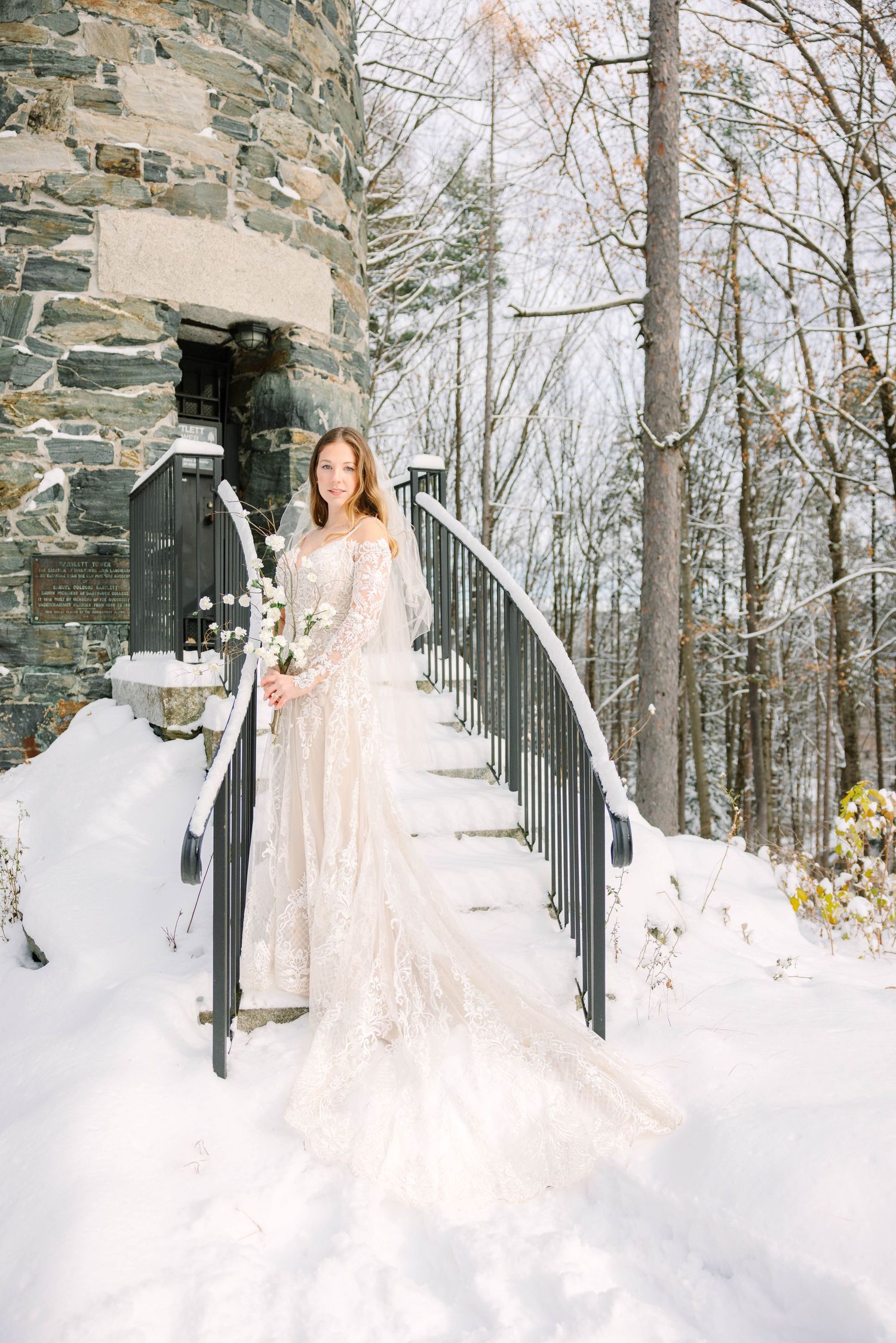 Bride in lace gown with veil holds white bouquet on snowy outdoor staircase beside stone building in winter.
