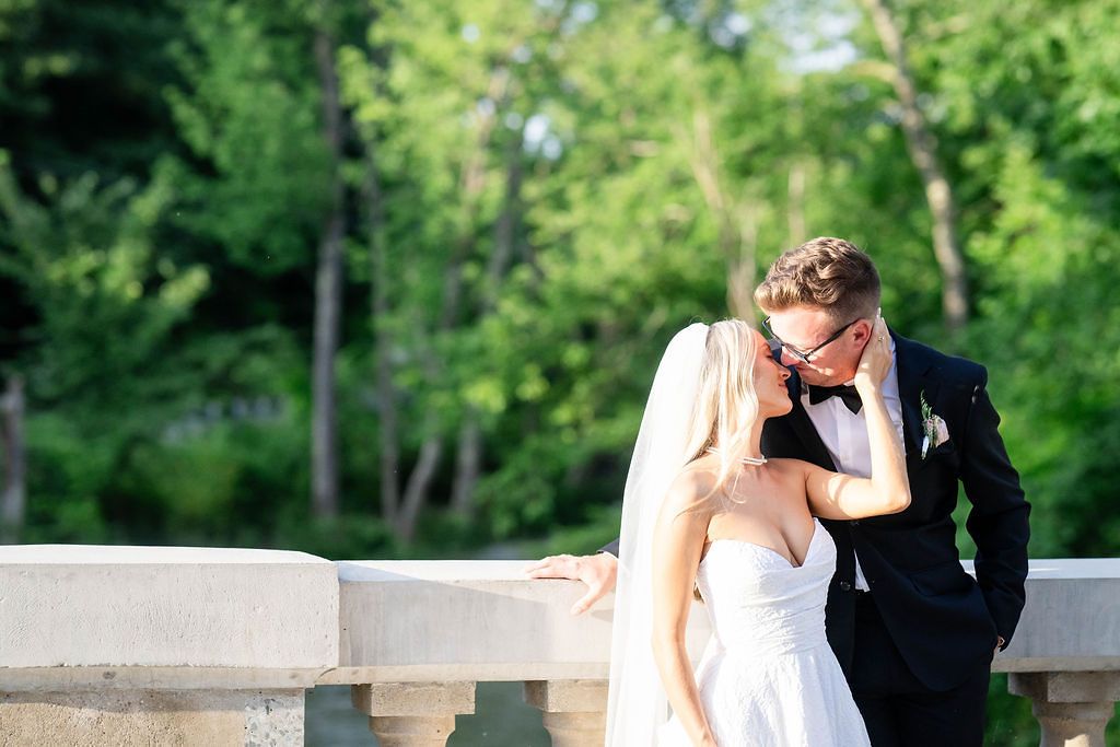 Bride and groom sharing a kiss on a stone balustrade bridge surrounded by lush green trees.