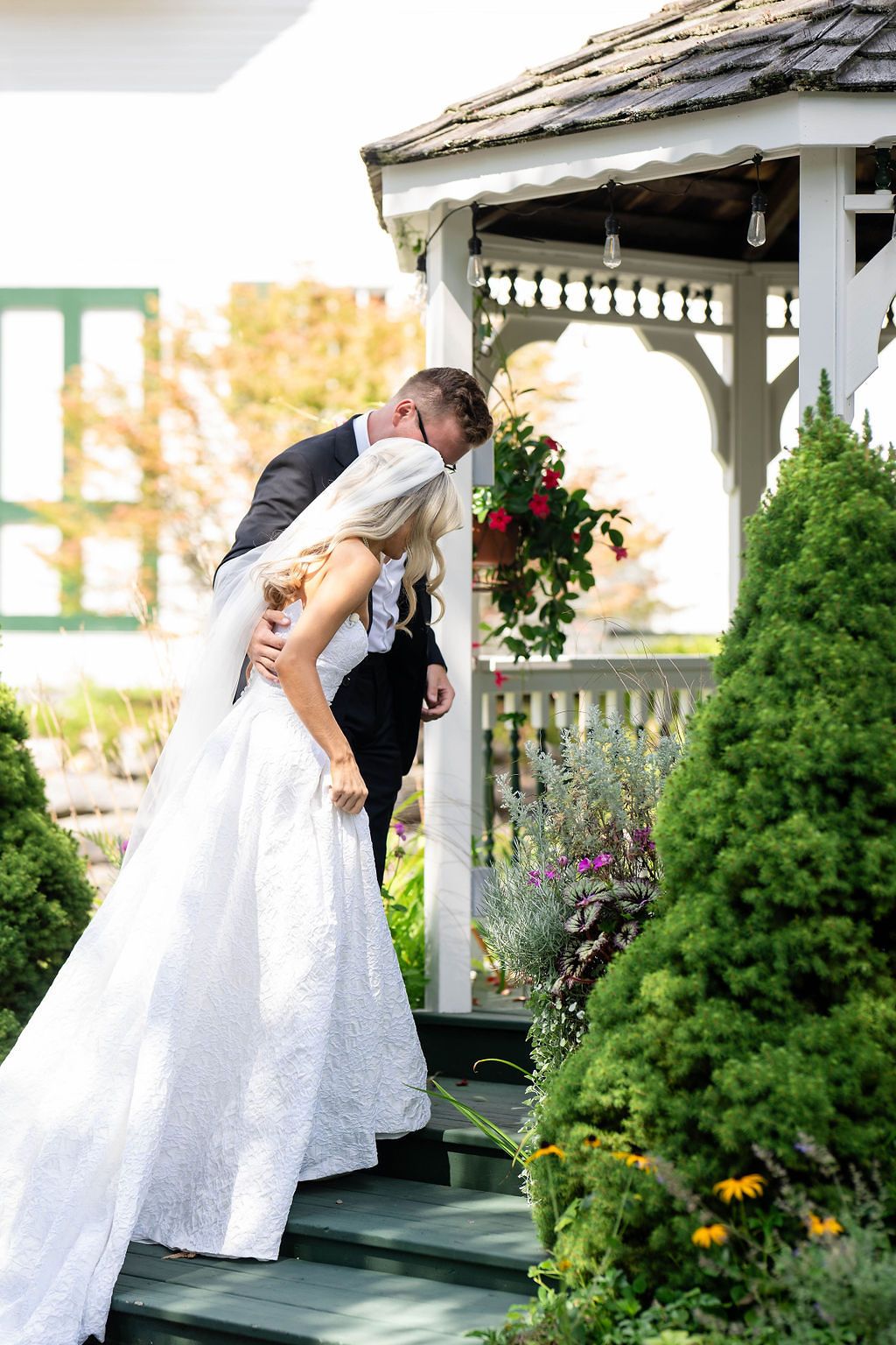 Bride and groom share a romantic kiss at a white gazebo adorned with red flowers and green shrubs.