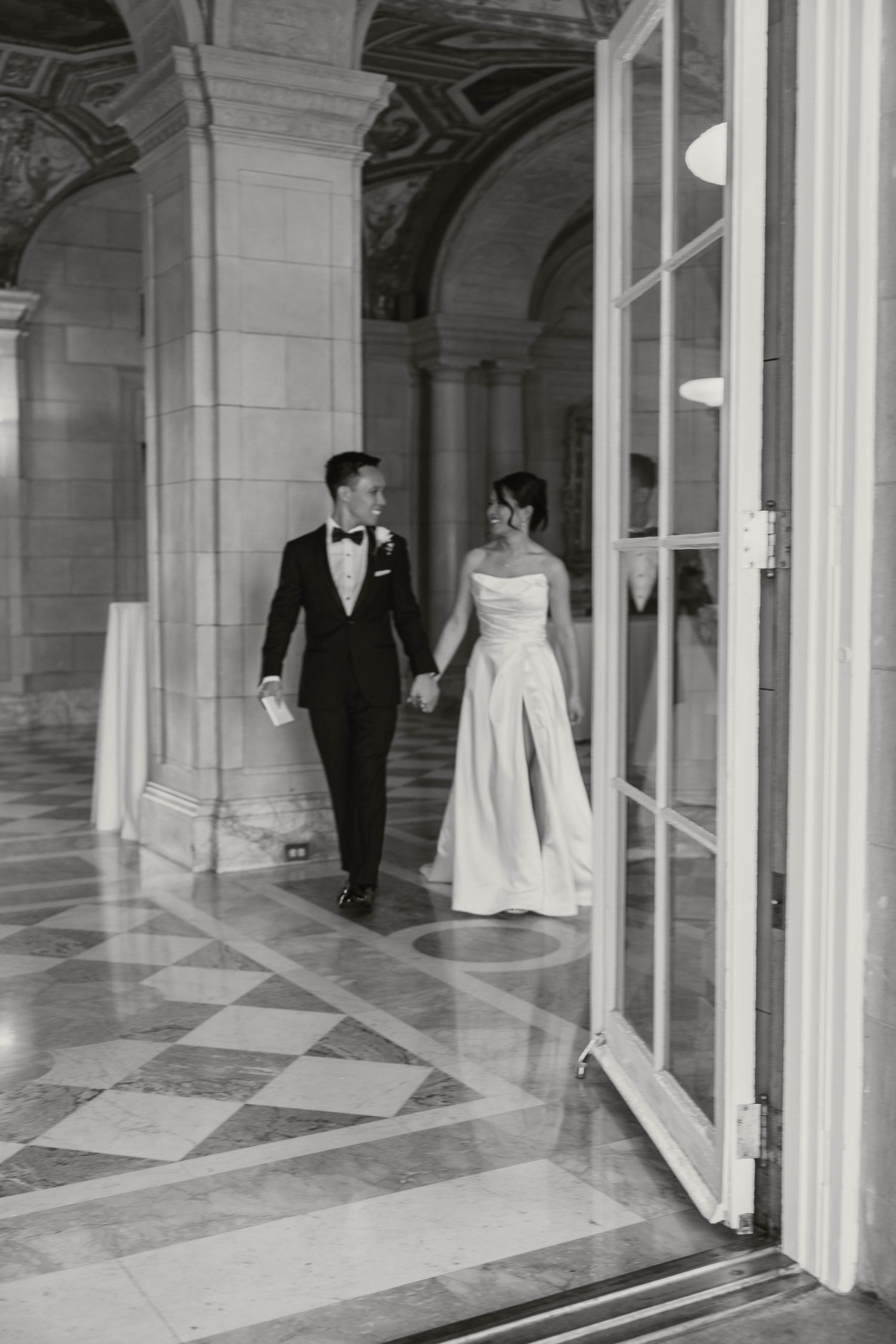 Bride and groom walk hand-in-hand through a grand marble-columned hall in black and white photo.