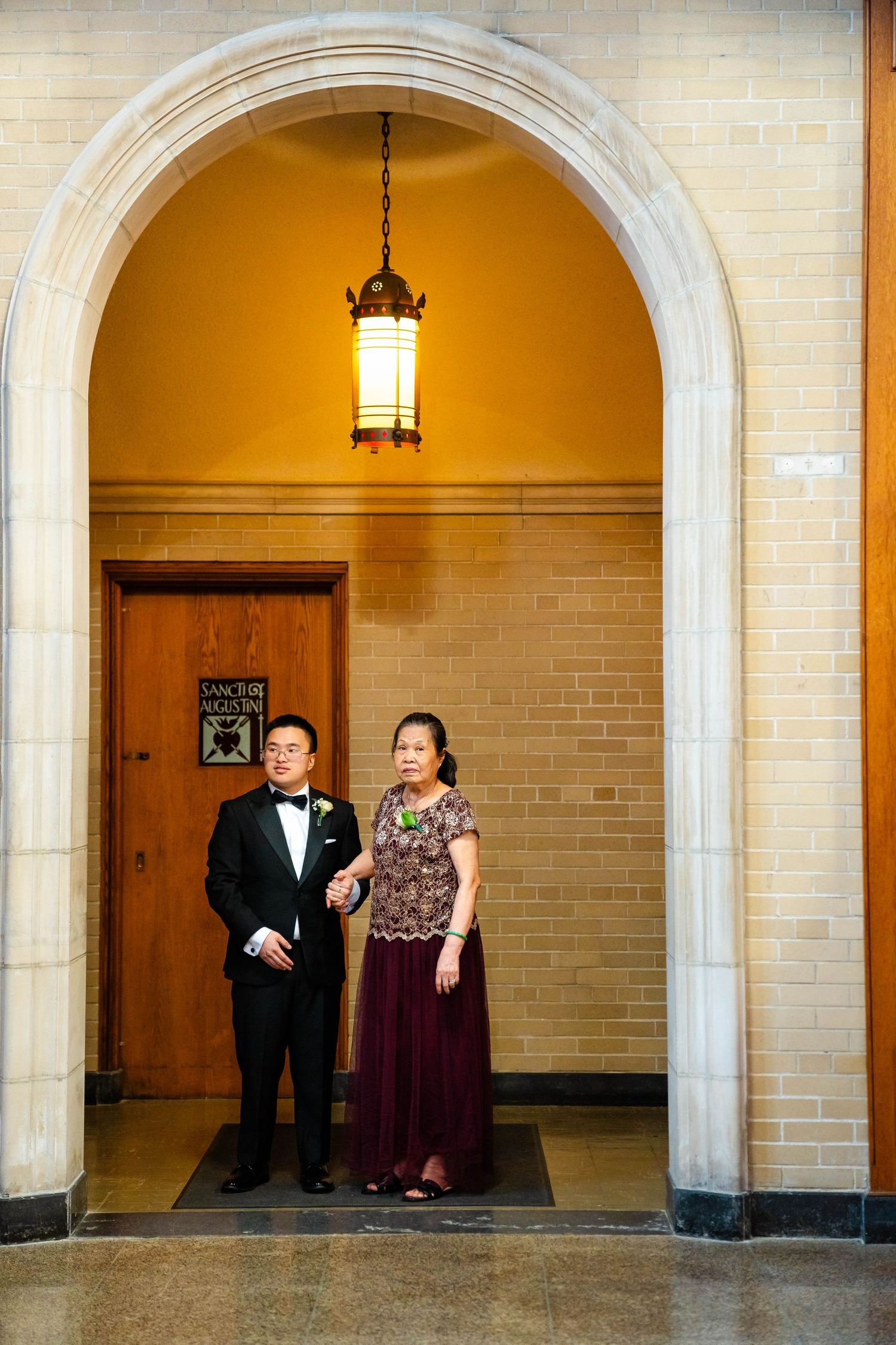Groom in tuxedo stands with mother in burgundy gown under ornate church archway.
