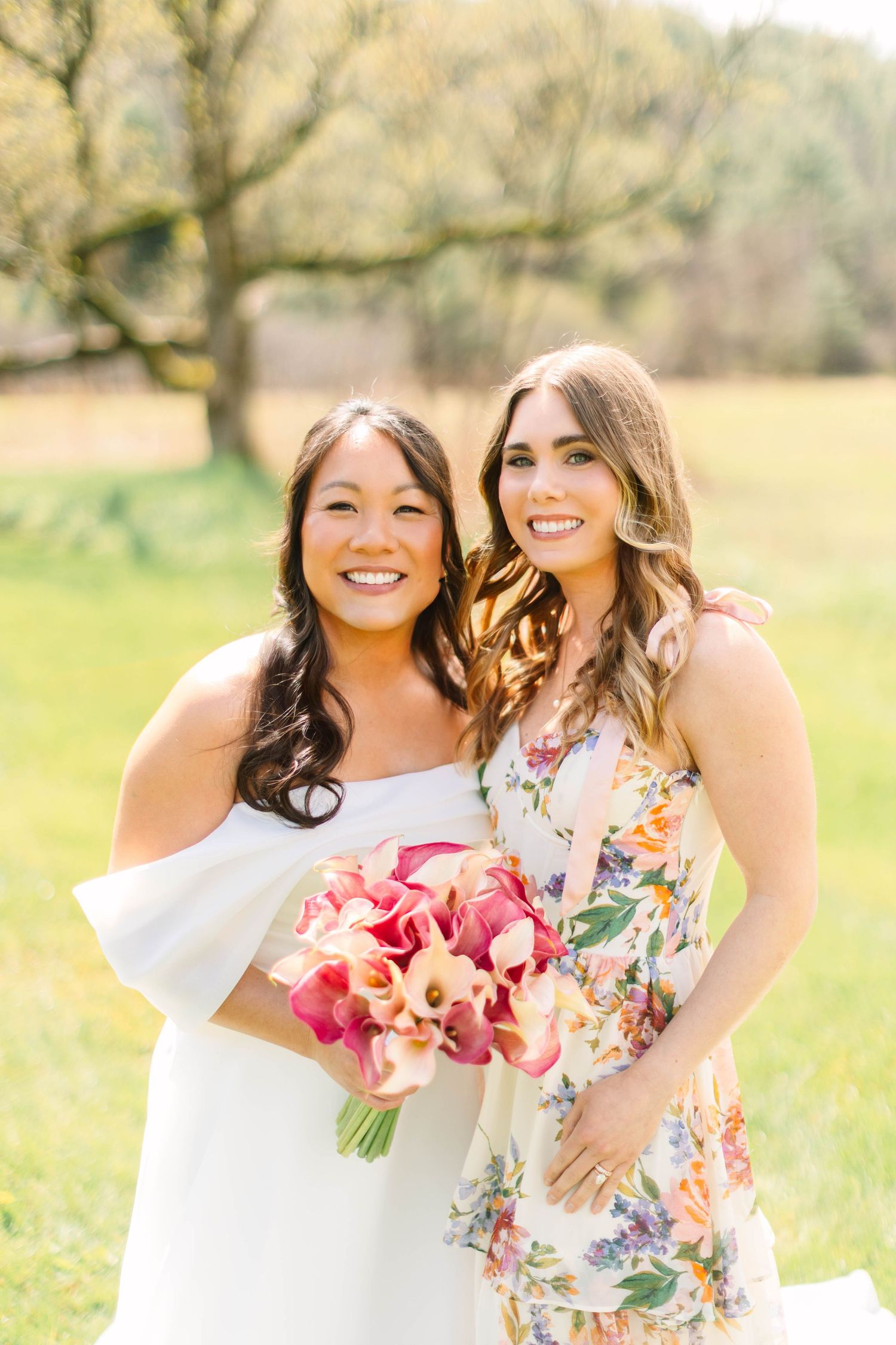Smiling bride in off-shoulder white gown holds pink calla lily bouquet with bridesmaid in floral dress.