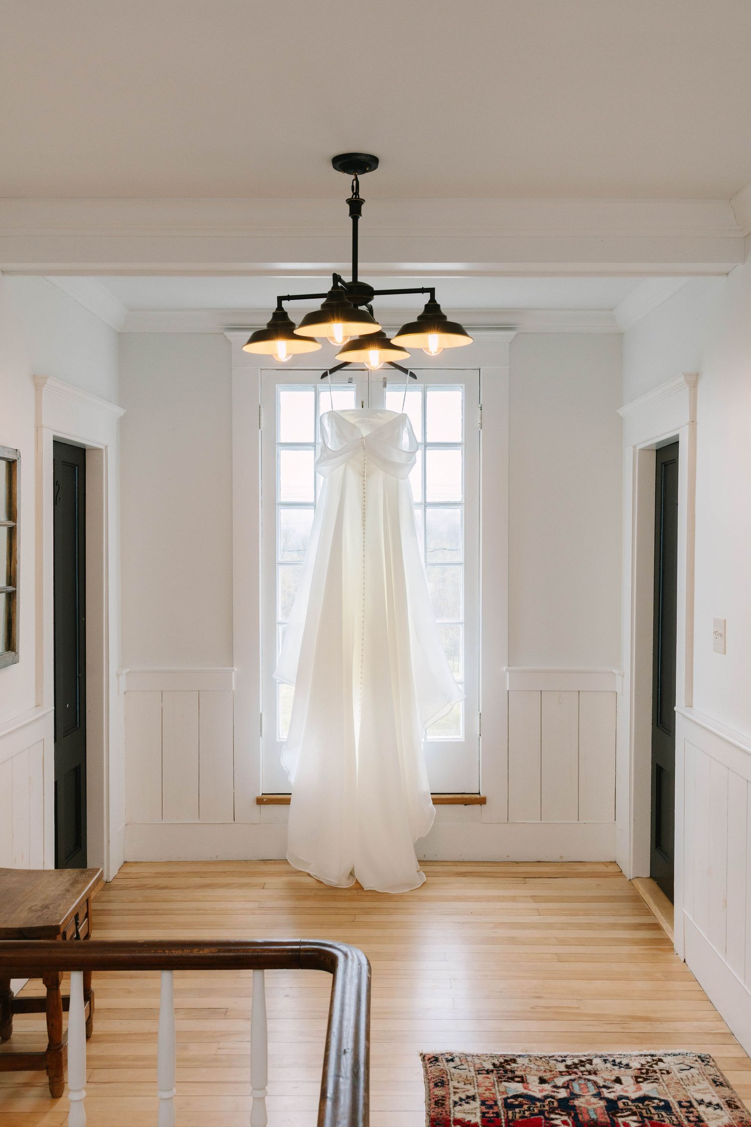 Elegant white wedding gown hanging from a black chandelier in front of a bright window.