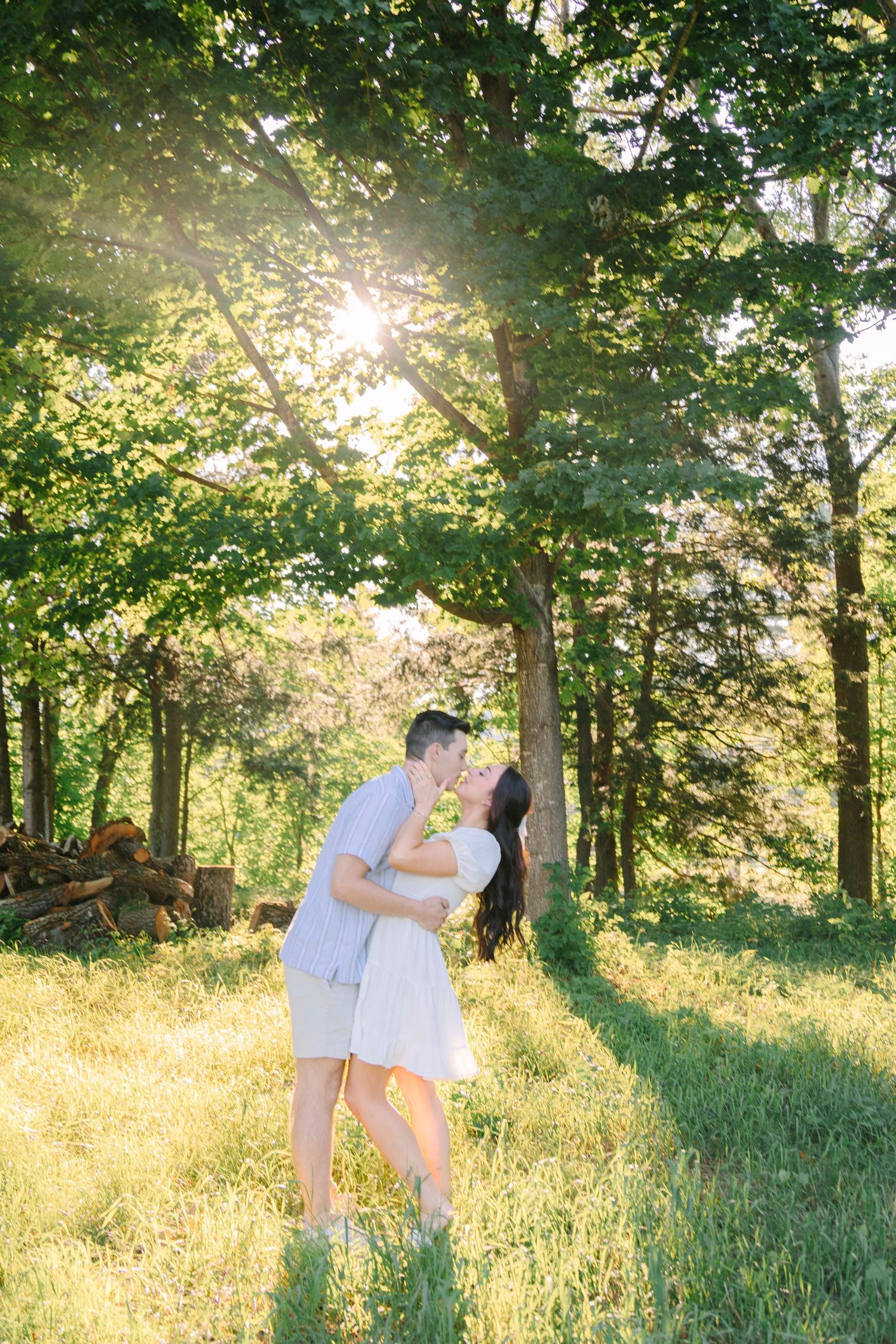 Couple sharing a romantic kiss in a sunlit forest clearing, surrounded by lush green trees on a warm summer day.