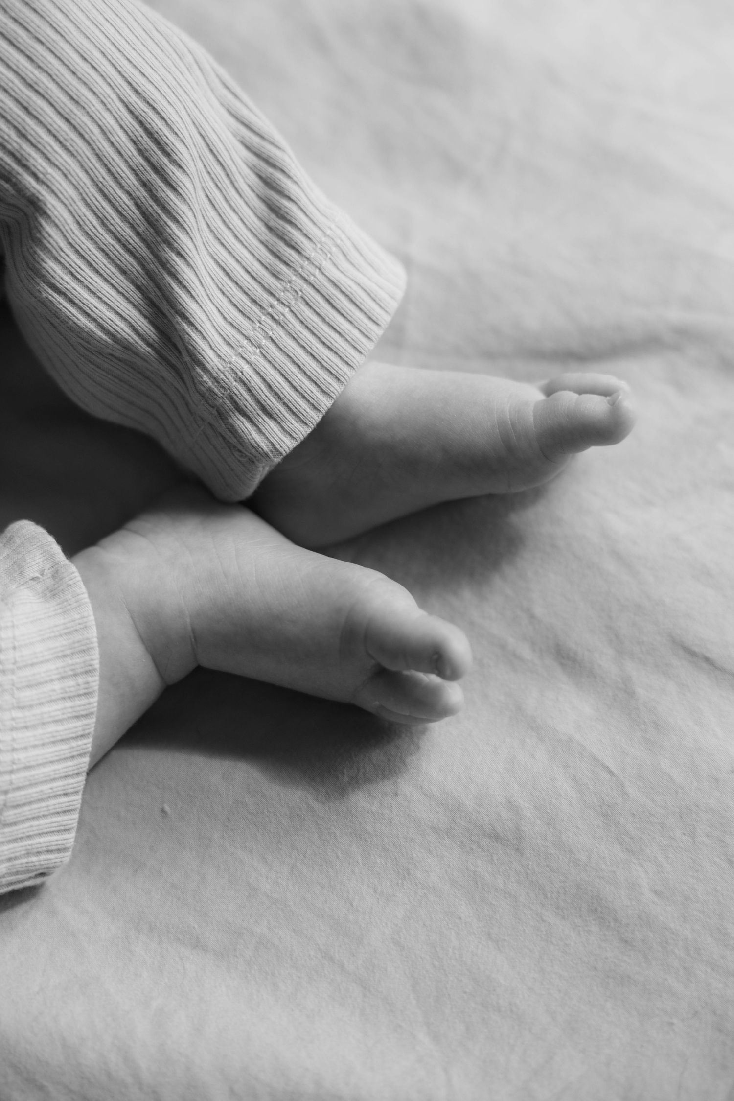 Close-up black and white photo of newborn baby feet in ribbed onesie on soft linen.