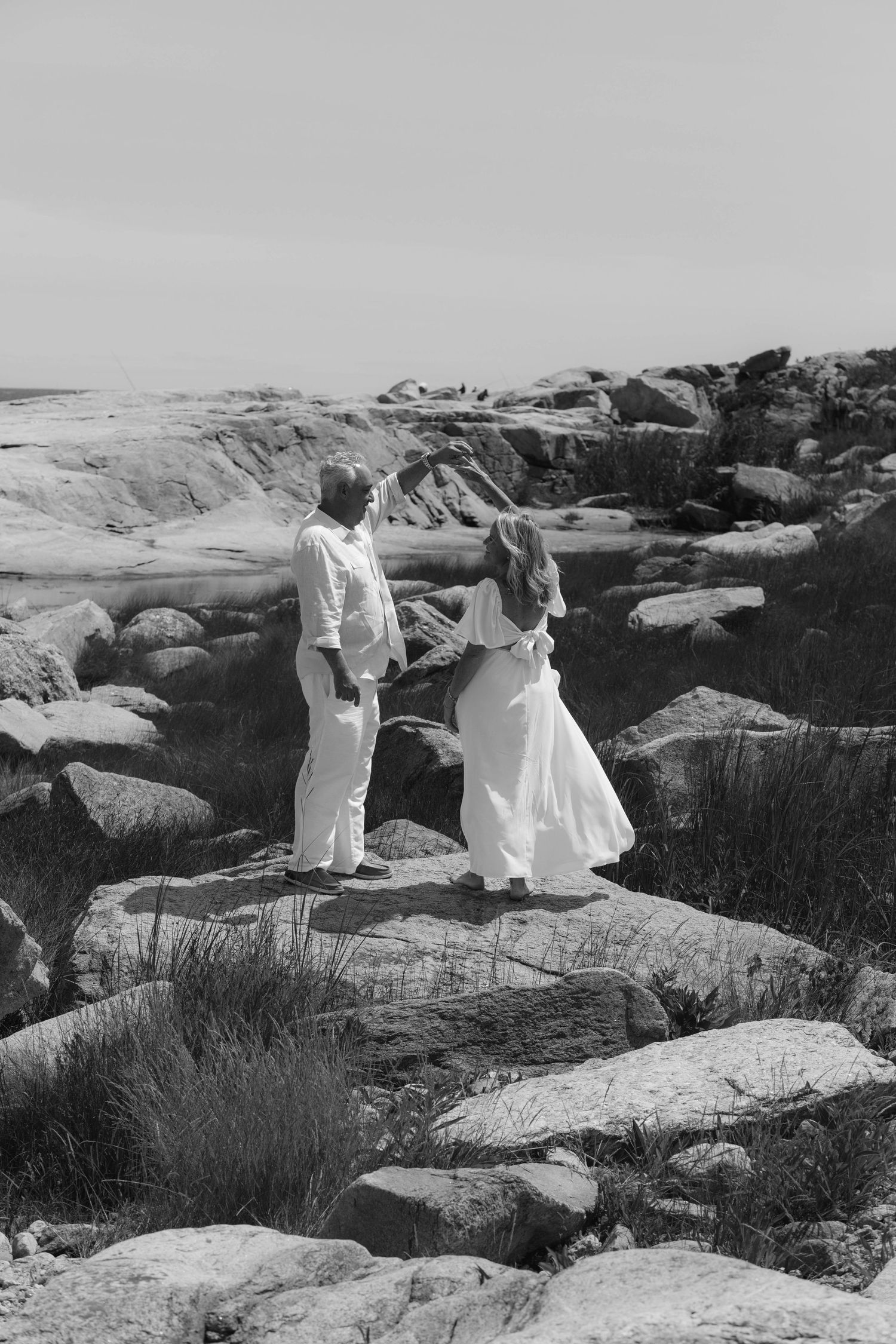 Black and white photo of a couple in white attire standing on rocky coastal terrain, facing each other romantically.