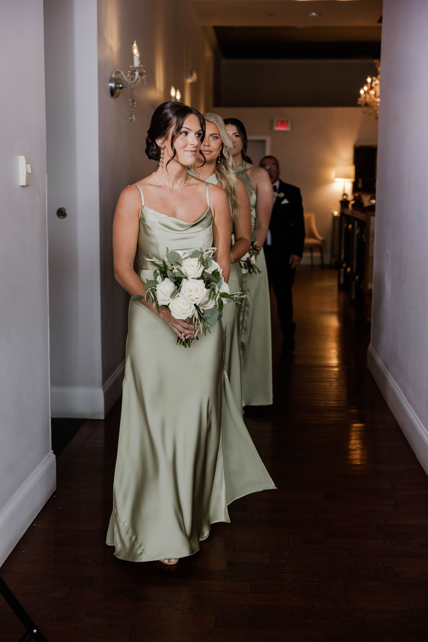 Bridesmaids in sage green satin gowns walk down a hallway carrying white floral bouquets at a wedding.