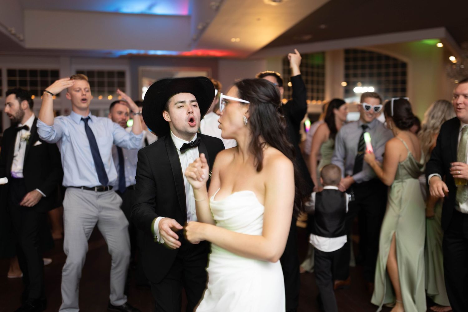 Bride and guest in cowboy hat dancing energetically on crowded wedding reception dance floor.