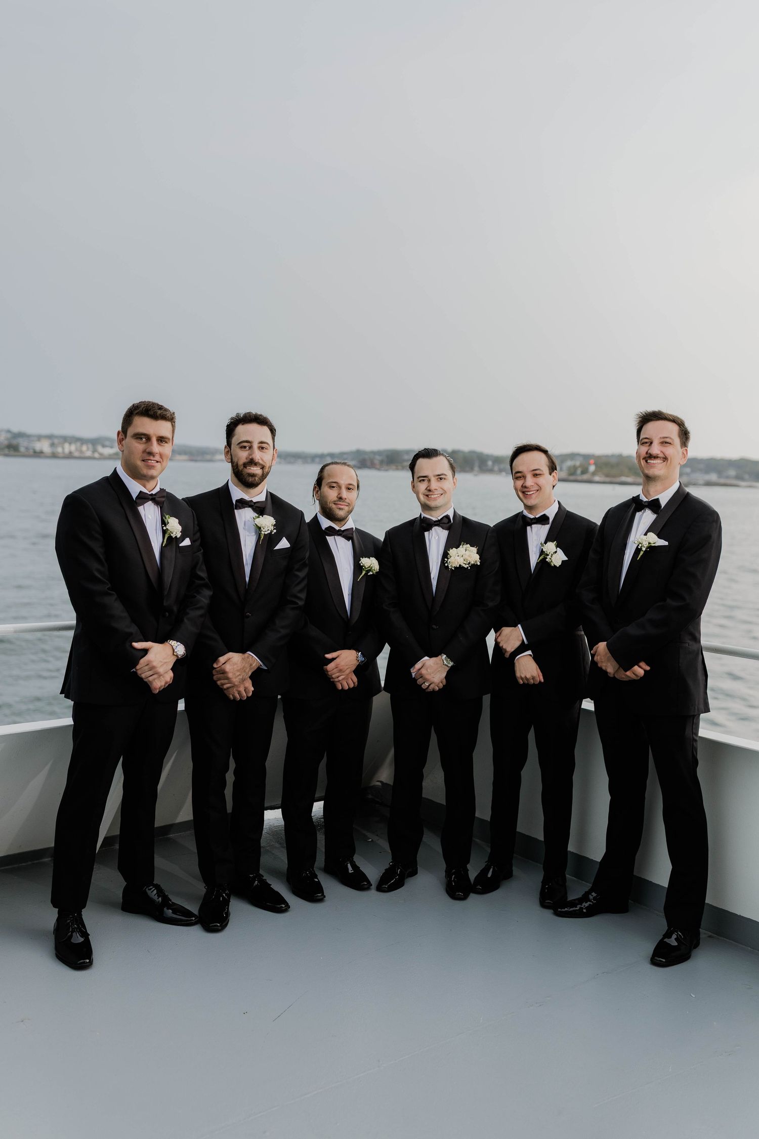 Groom and five groomsmen in black tuxedos pose together on a boat deck with water views.