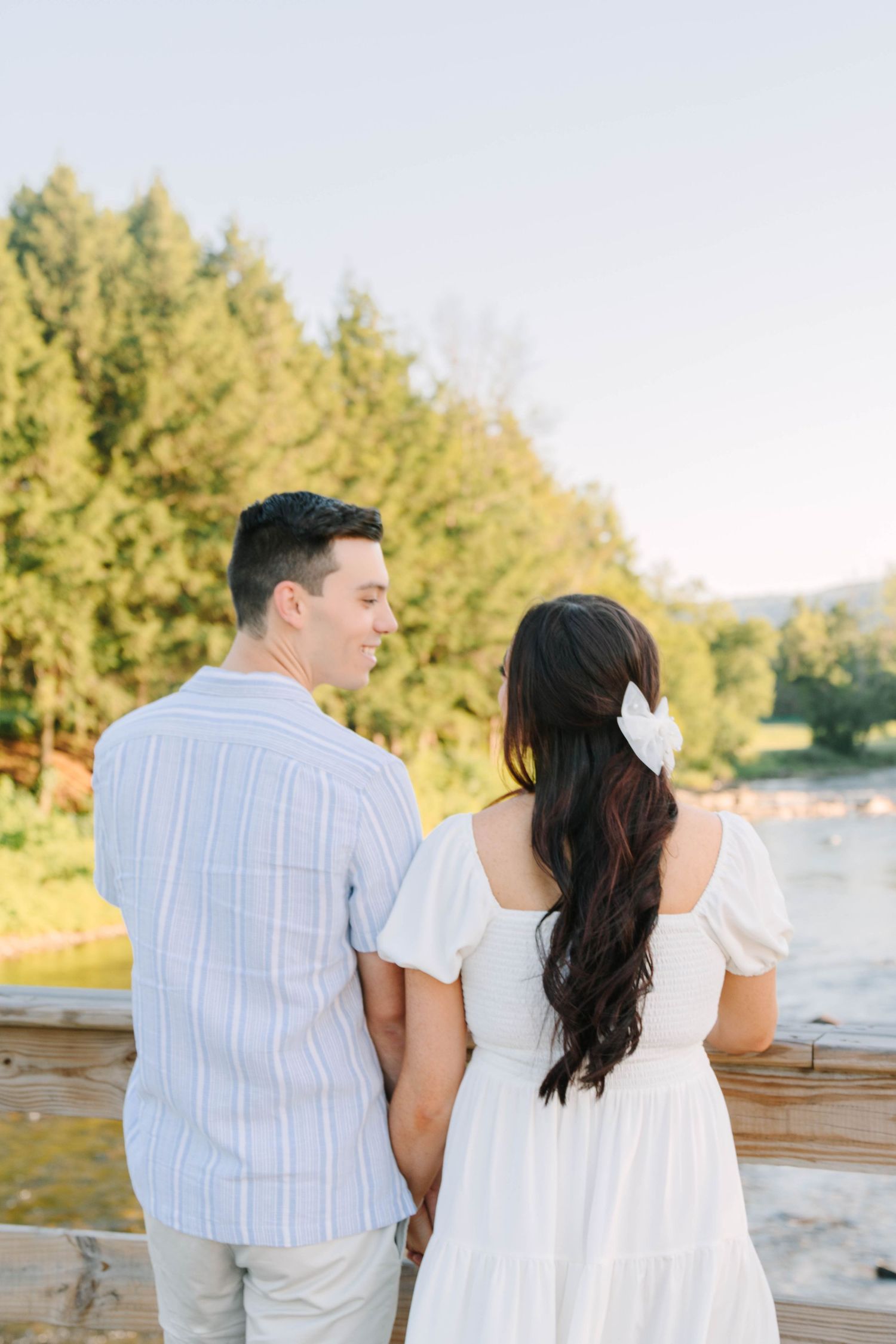 Couple holding hands on a wooden bridge overlooking a river surrounded by lush green trees at golden hour.