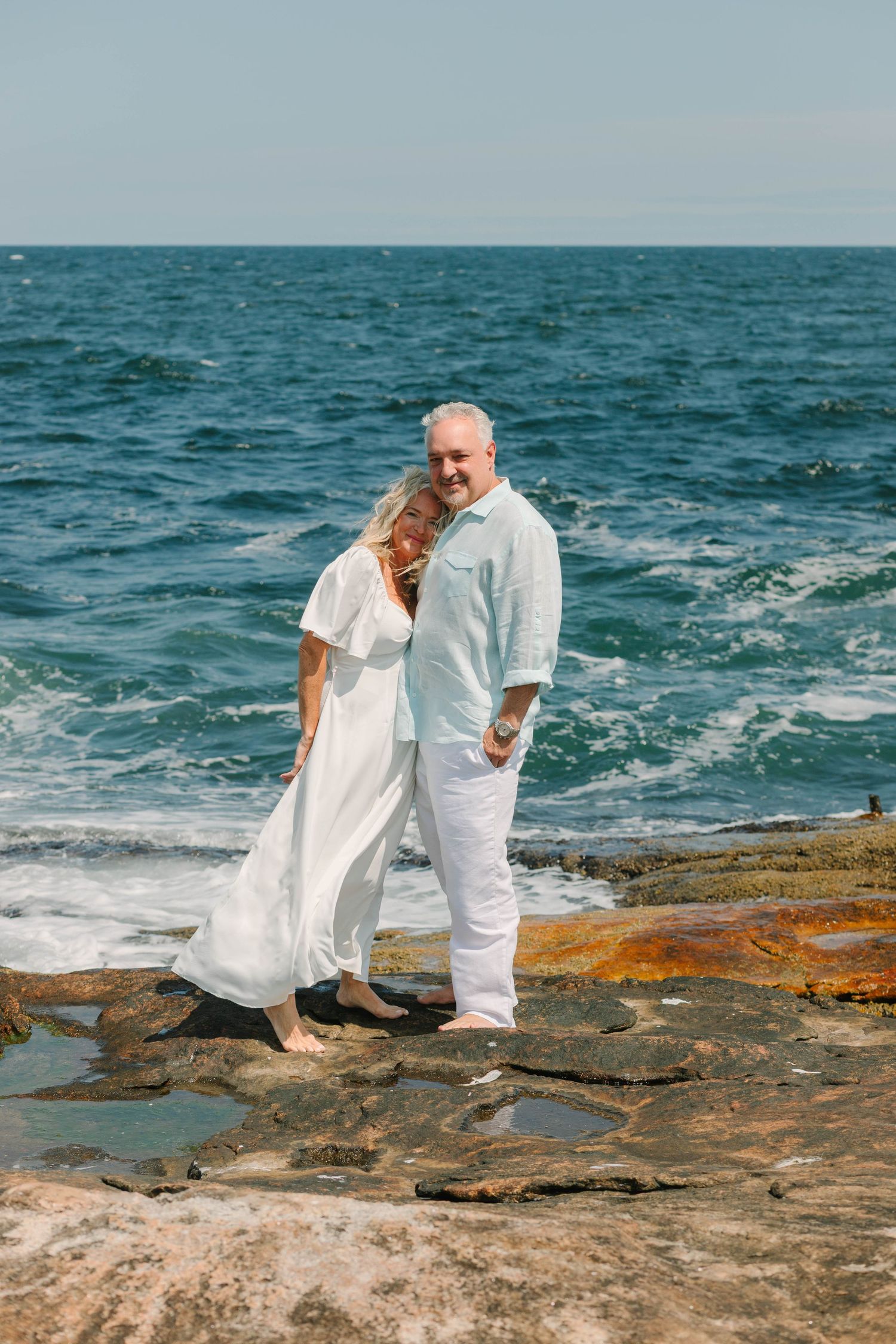 Mature couple in white clothing standing on rocky coastline with blue ocean waves in background.