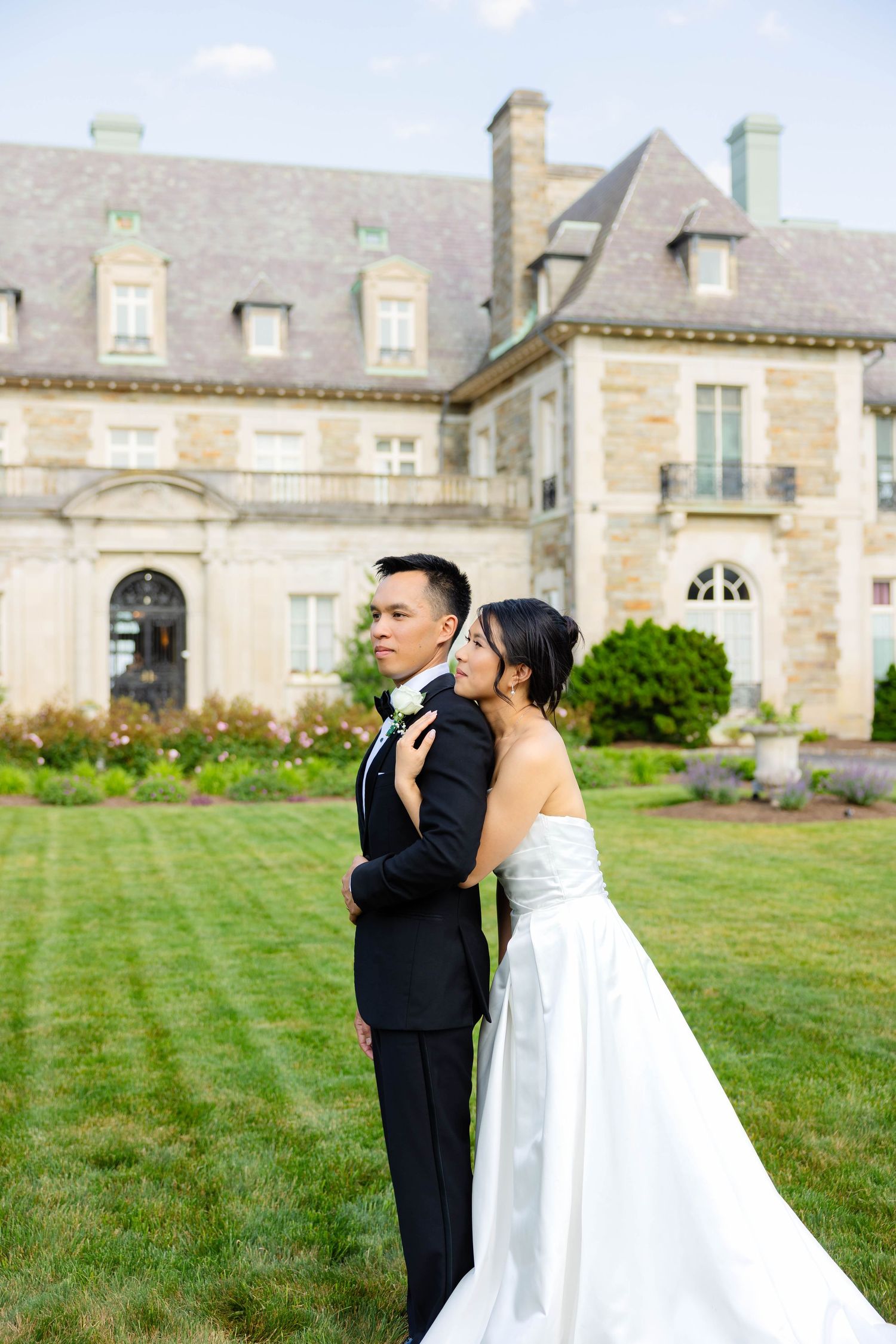 Bride embracing groom from behind in front of grand stone estate on wedding day.