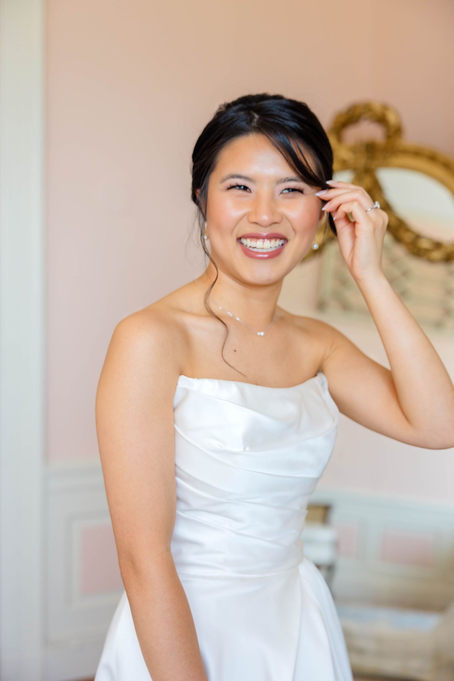 Smiling bride in strapless white wedding gown adjusting her earring in an elegant room.