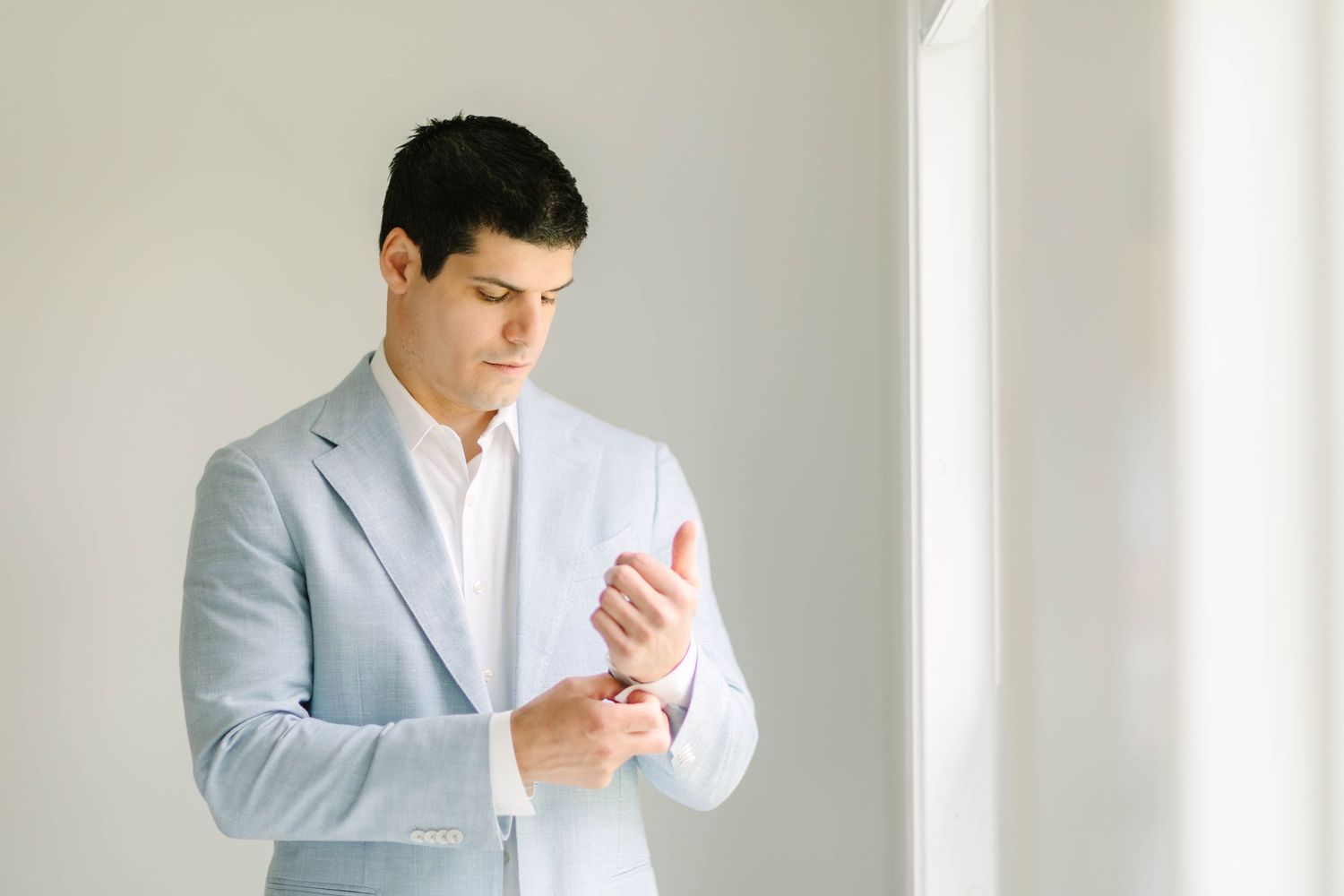 A well-dressed man in a light blue suit adjusts his cufflinks while standing near a bright window.