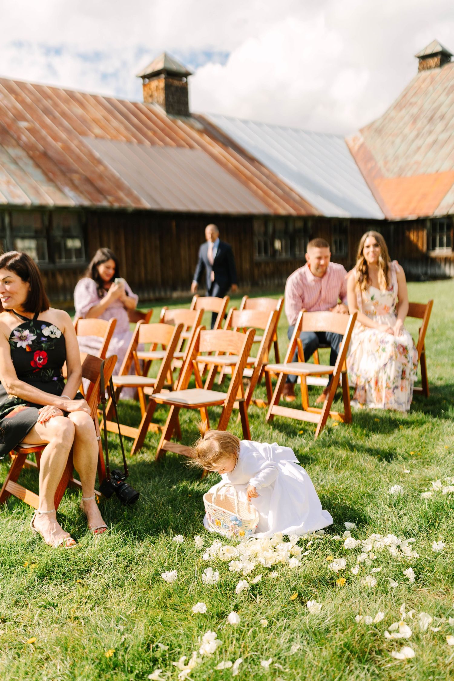 Adorable flower girl scatters white petals on grass aisle at outdoor barn wedding ceremony.