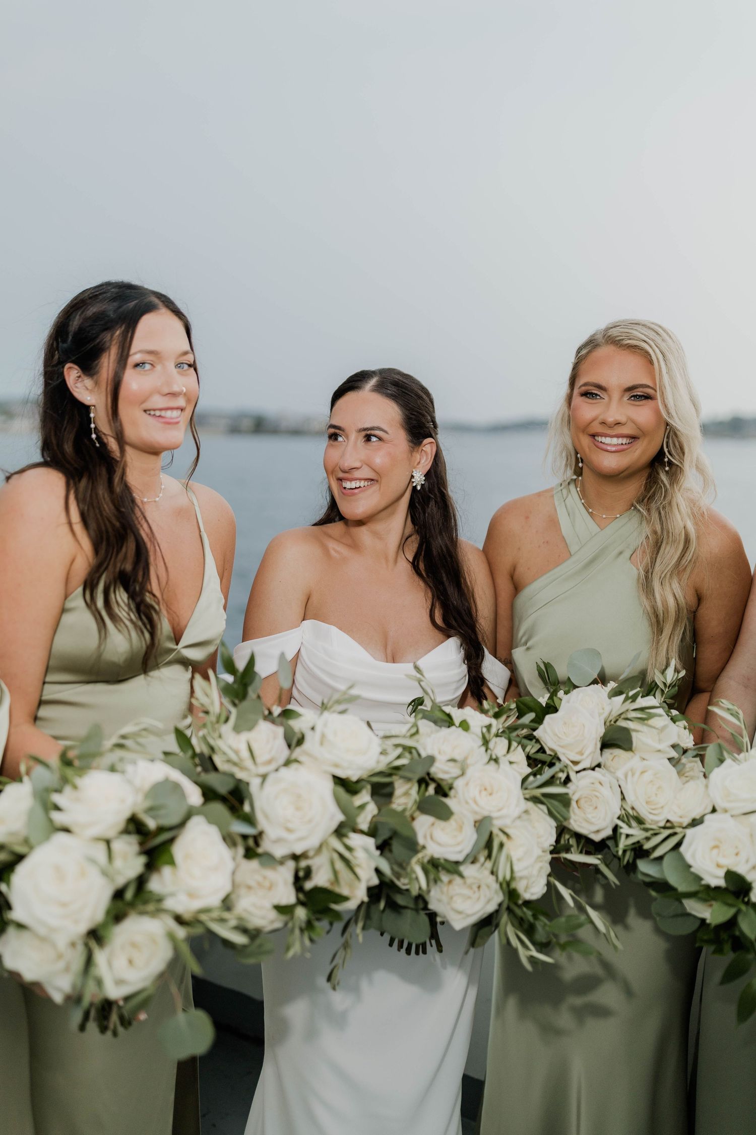 Bride poses with bridesmaids in sage green gowns holding white rose bouquets waterside.