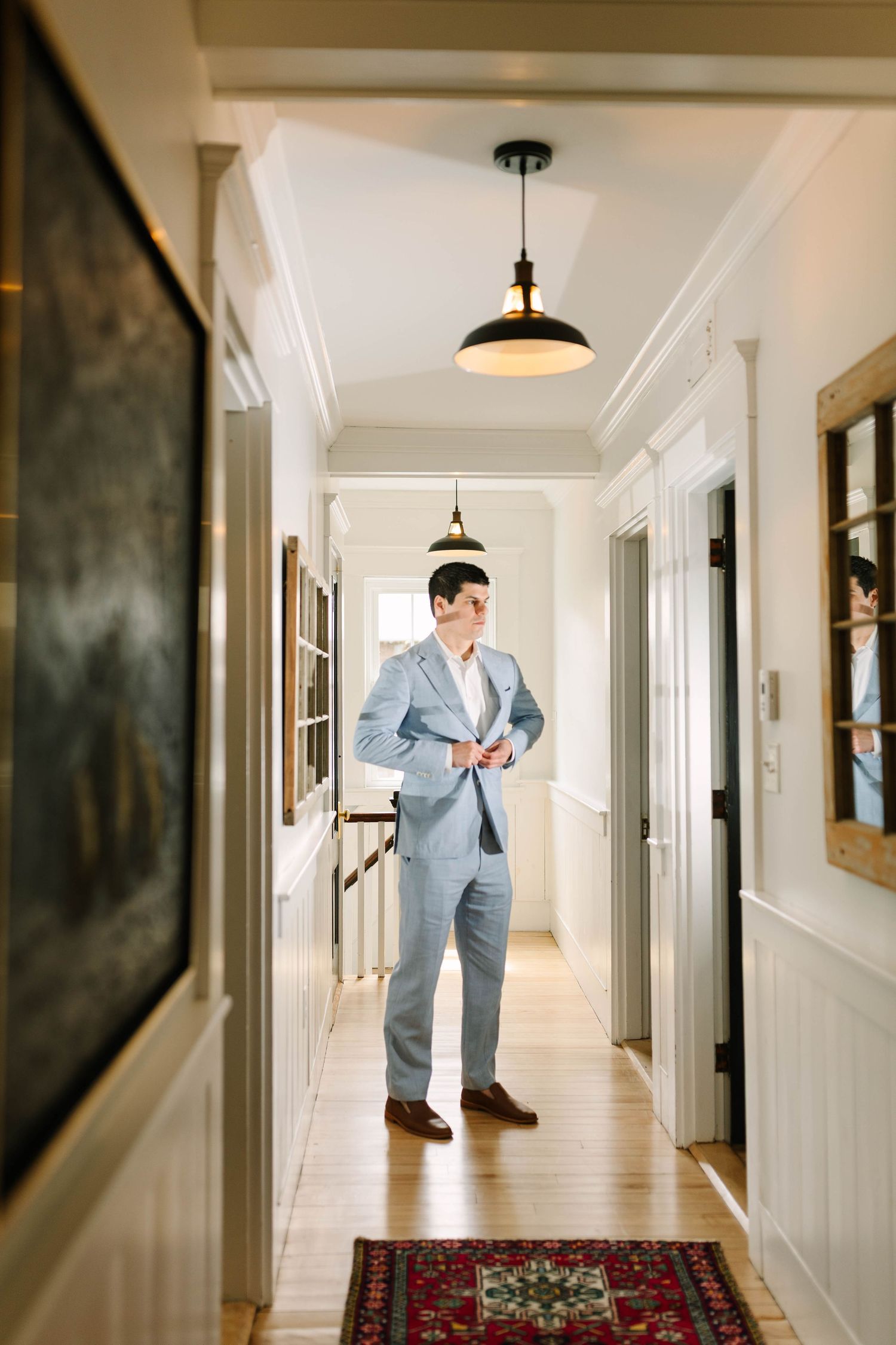 Groom in light blue suit buttoning jacket while standing in elegant white hallway with hardwood floors and Persian rug.