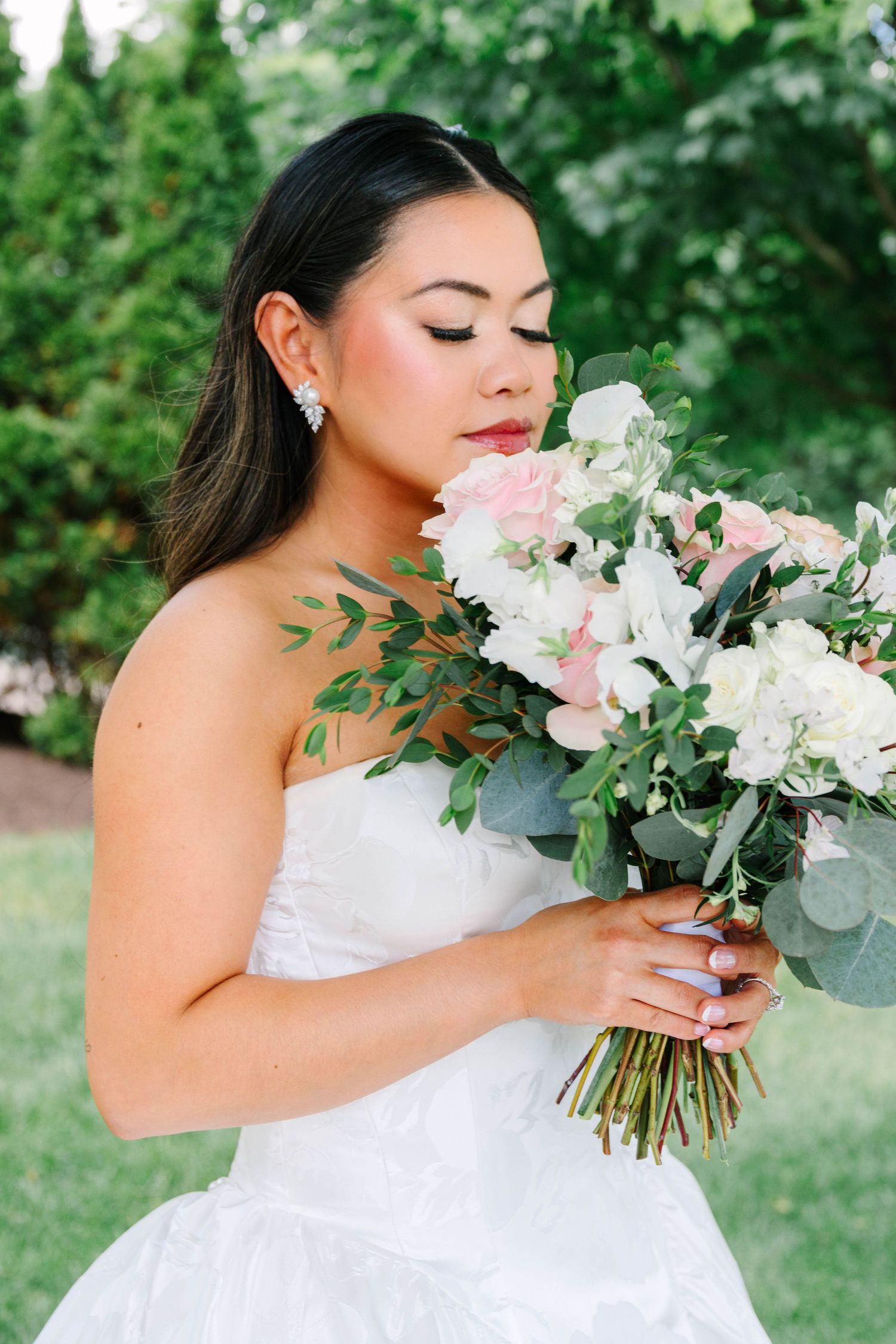Beautiful bride in strapless white gown smelling a pink and white floral bouquet outdoors.