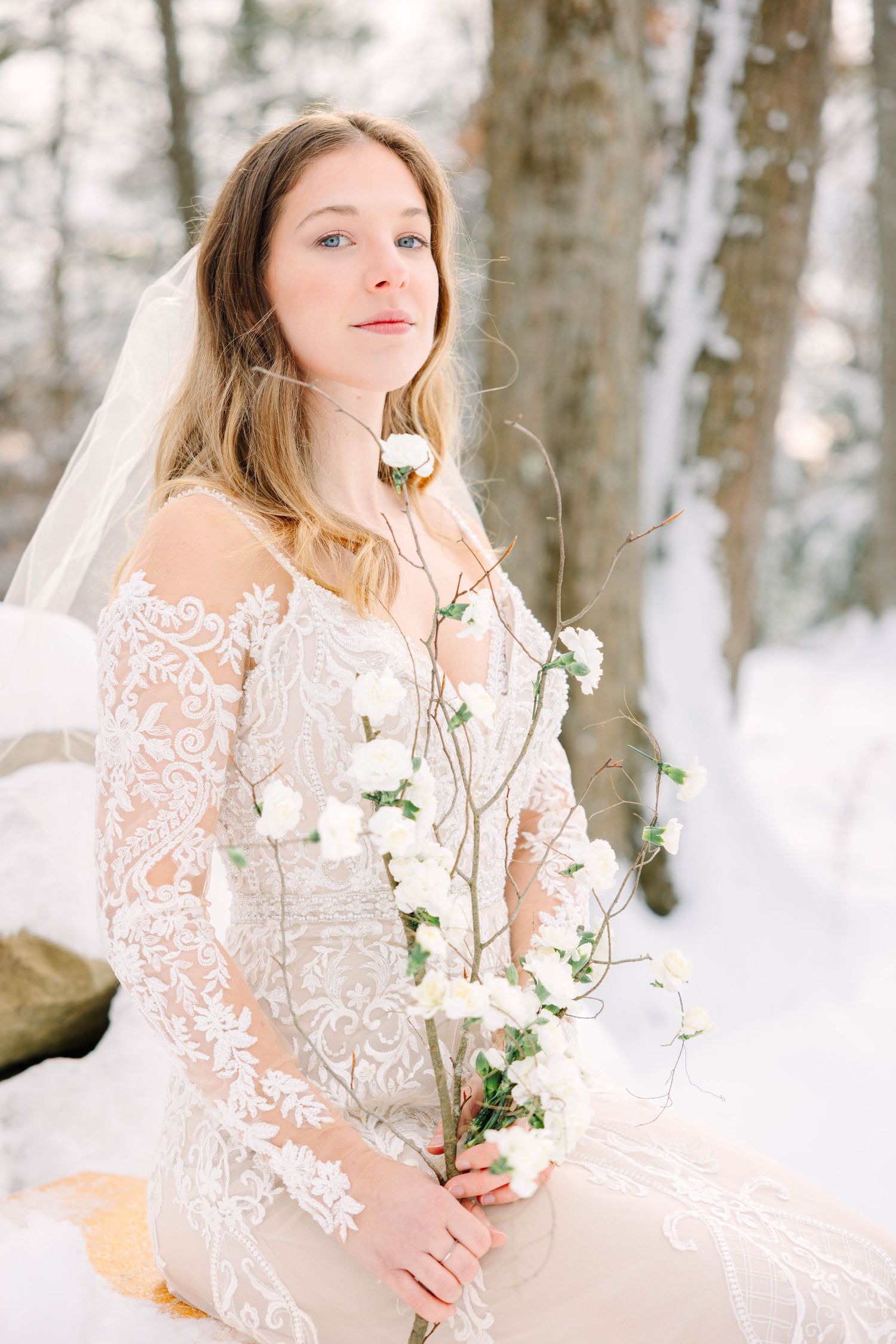 Serene bride in lace gown with veil holding white floral branch in snowy winter forest.
