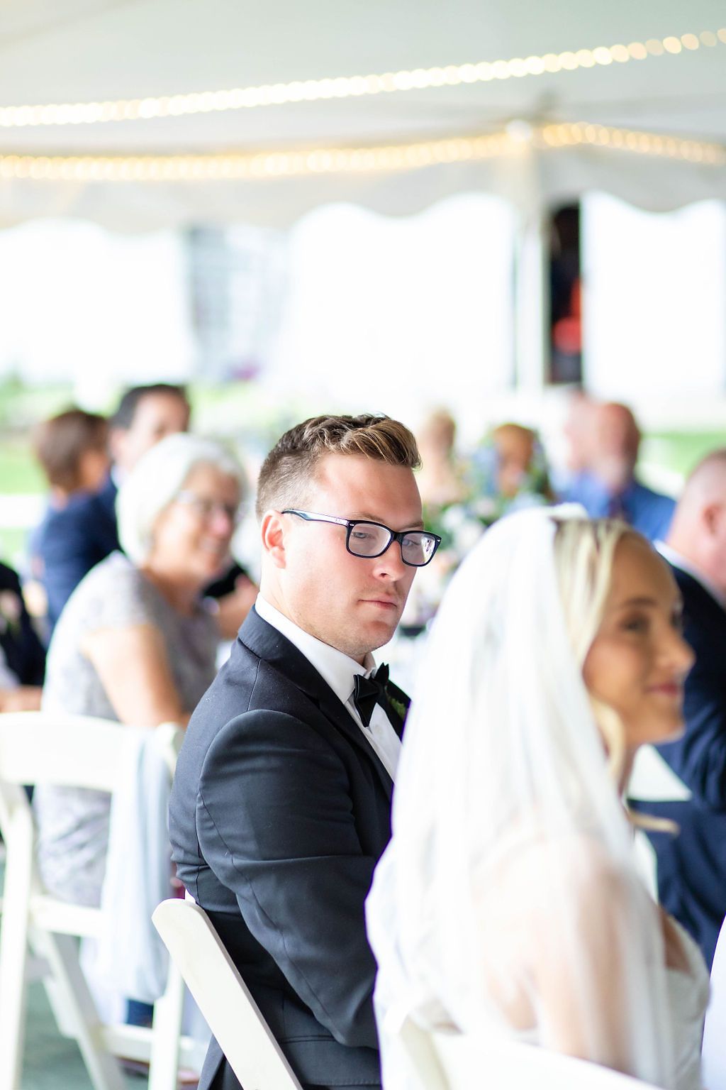 Groom in black tuxedo with glasses looks back during wedding reception under a white tent.