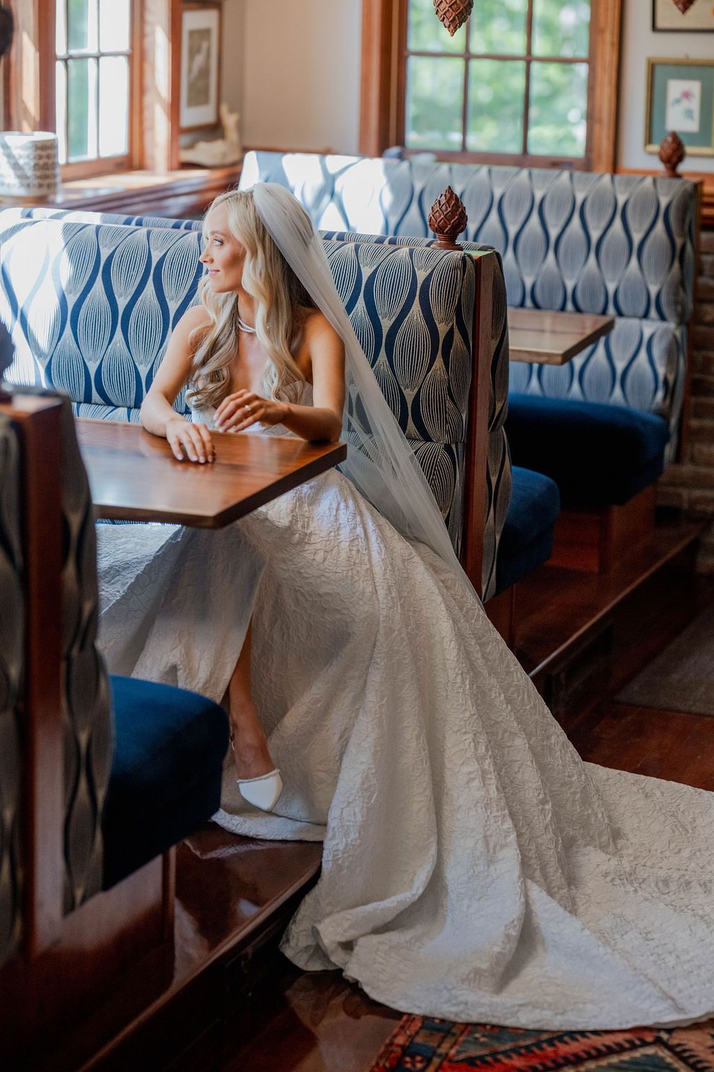 Bride in lace wedding gown and veil sits at wooden booth with blue patterned upholstery in elegant venue.