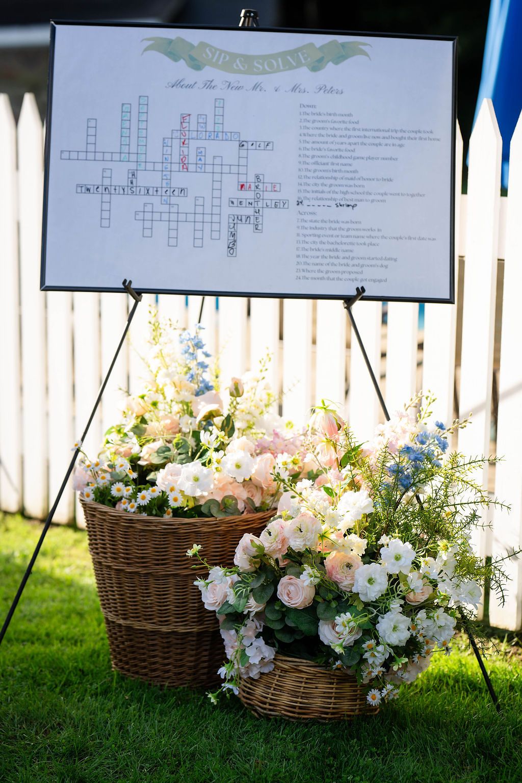 Floral basket arrangement with pink and white flowers beside a crossword puzzle wedding sign on an easel outdoors.