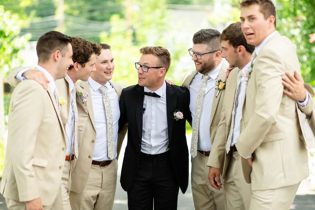 Groom in black tuxedo laughing with groomsmen in tan suits at outdoor wedding celebration.