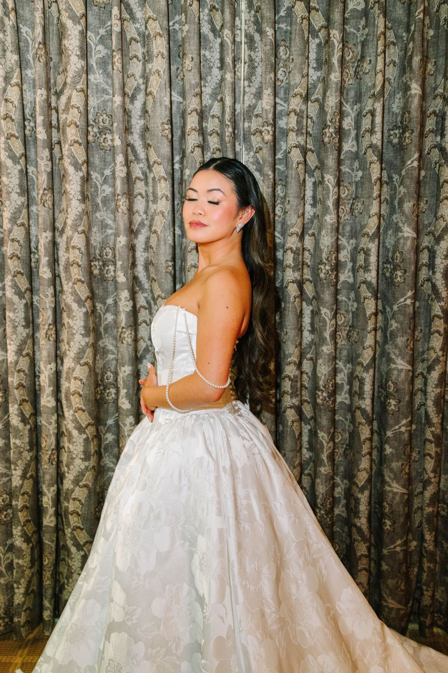 Bride in strapless white floral ballgown with eyes closed posing before gray curtain backdrop.