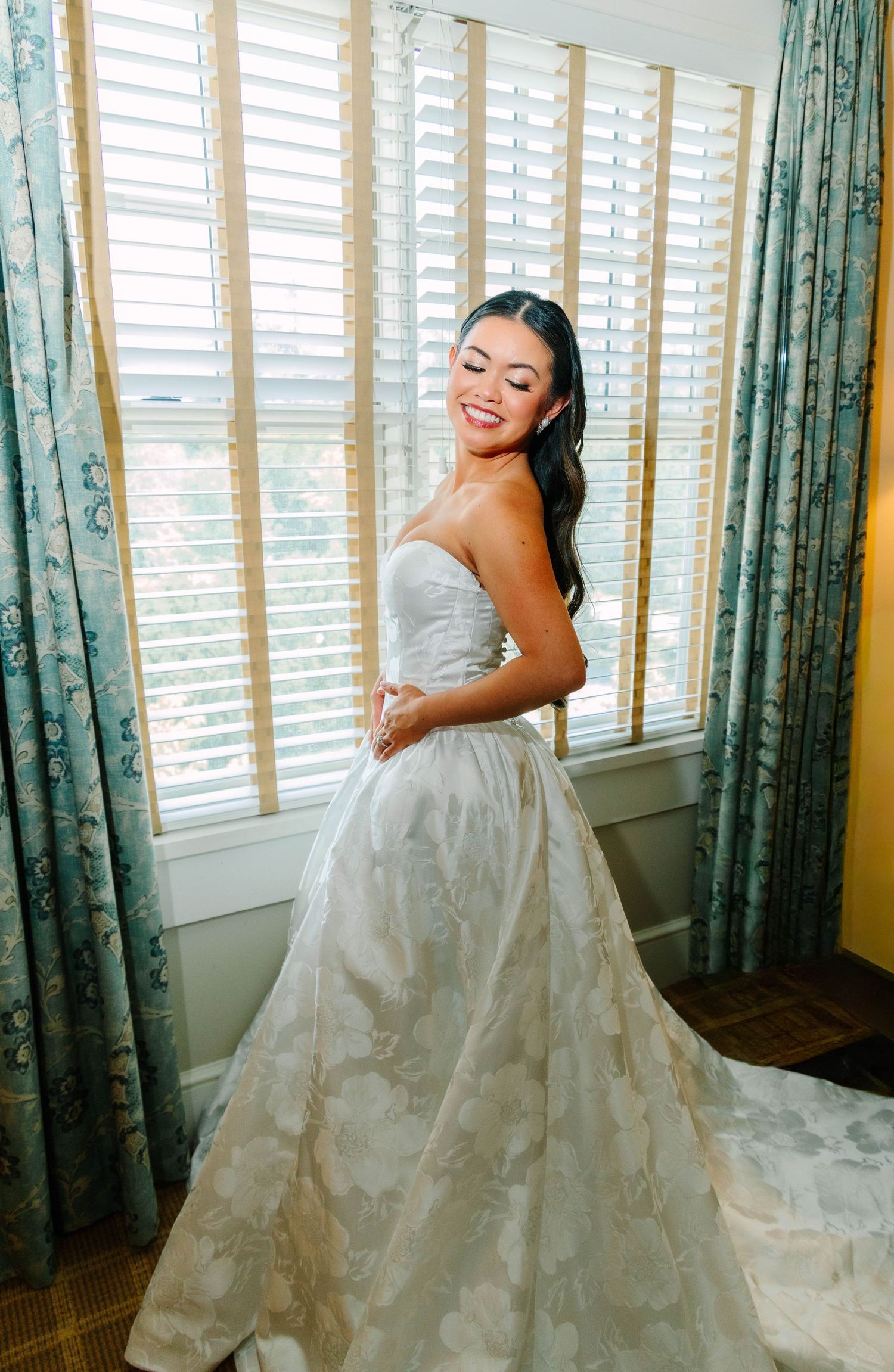 Smiling bride in a strapless ivory floral ballgown standing by sunlit window with teal curtains.