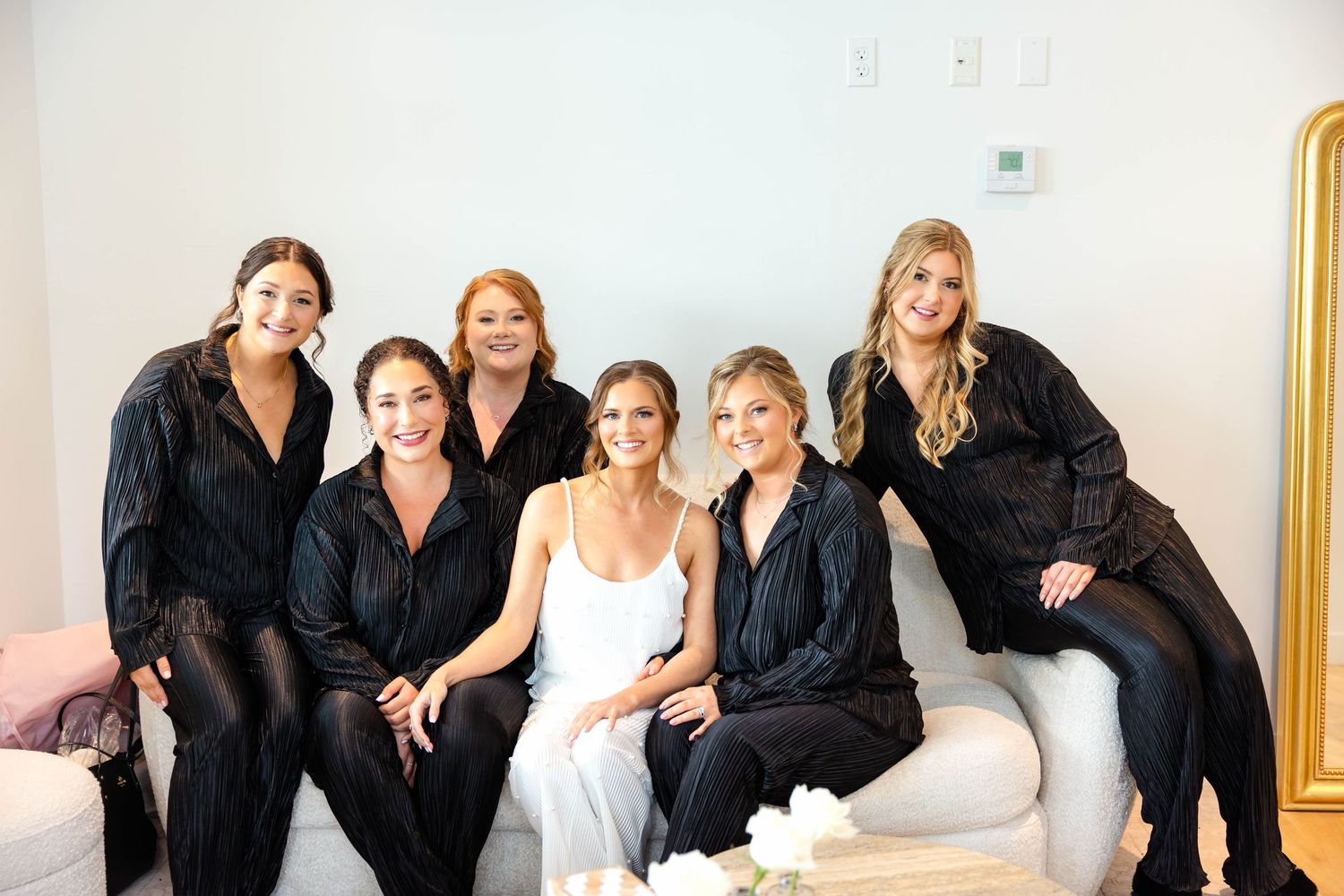 Bride surrounded by five bridesmaids in black satin robes smiling together in bright room.