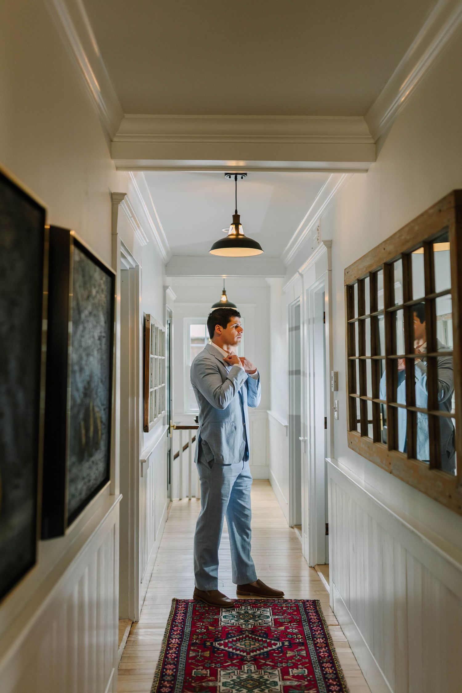 Groom in light blue suit adjusting tie in elegant white hallway with Persian rug and hardwood floors.