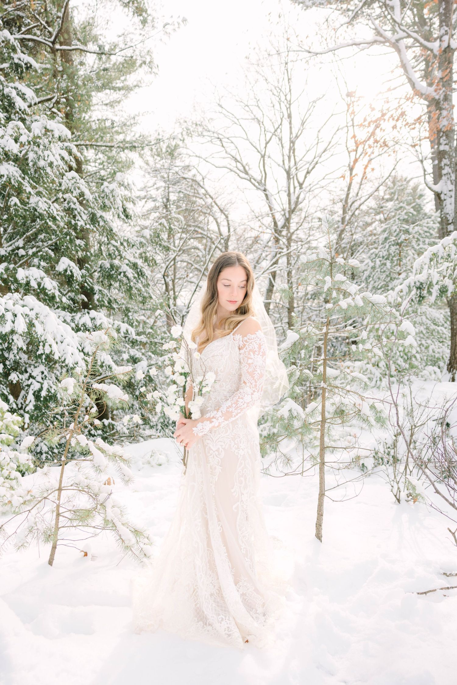 Bride in lace gown holding white flowers in snowy winter forest.
