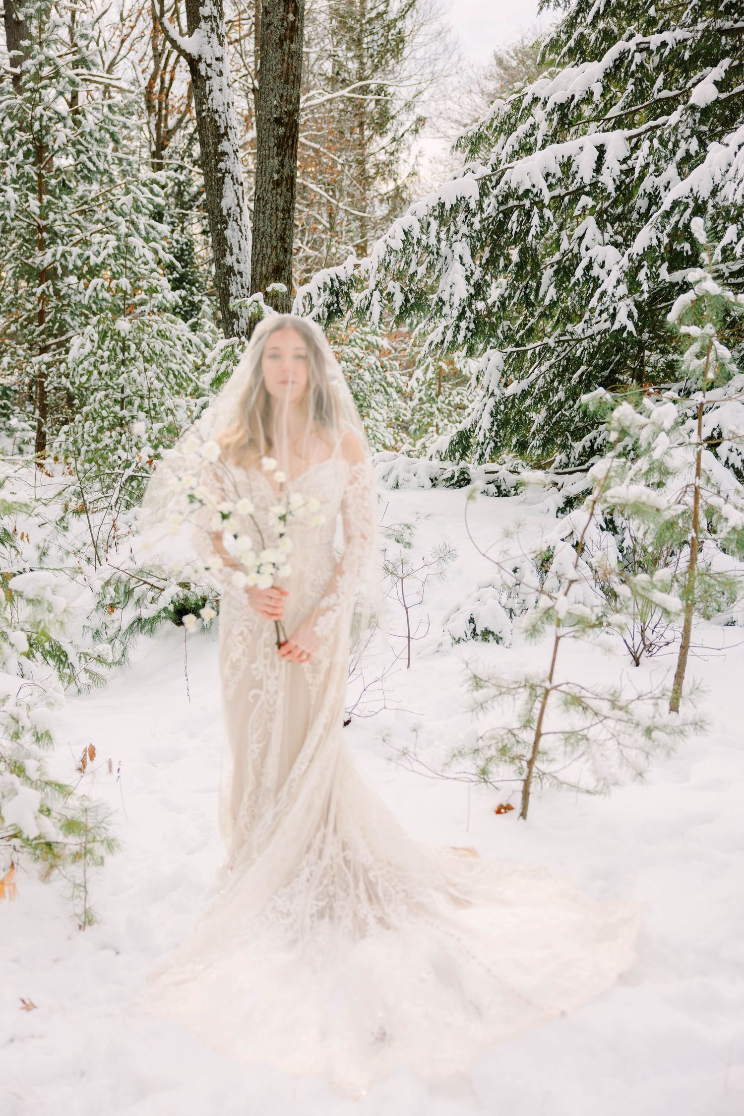 Full-length bride in lace gown and veil stands holding white blooms surrounded by snow-covered pines.