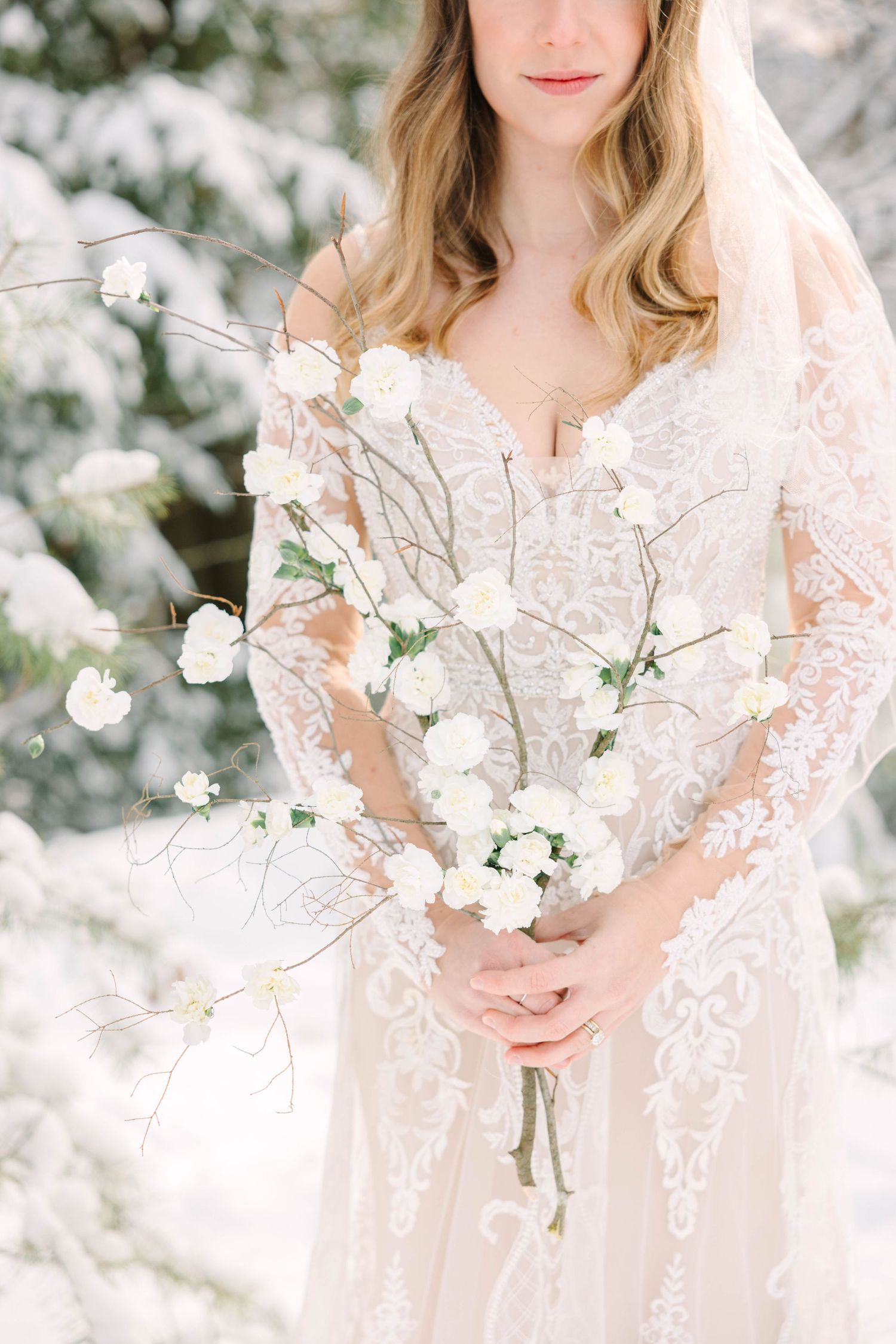 Bride in lace wedding gown holding delicate white floral branches in a snowy winter forest setting.