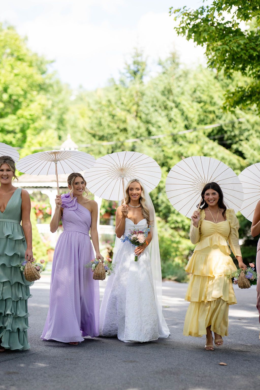 Bride in white lace gown walks with bridesmaids in pastel dresses holding white parasols in a garden.