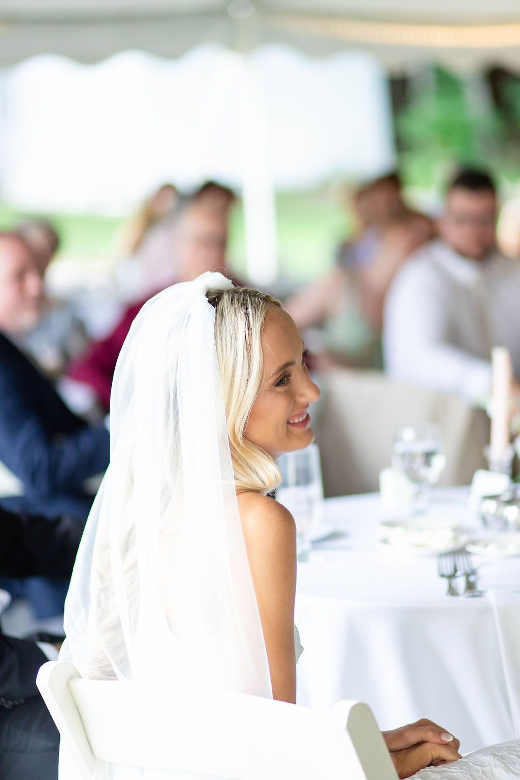 Smiling bride in white veil sits gracefully at wedding reception table surrounded by guests.