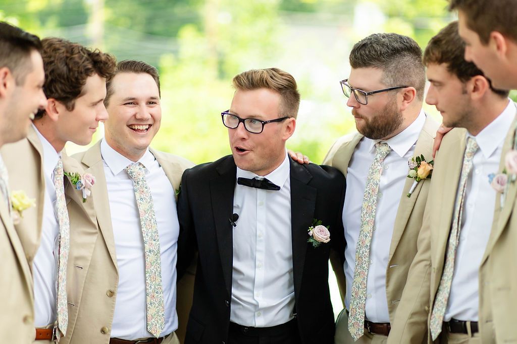 Groom in black tuxedo laughs with groomsmen wearing tan suits and floral ties at wedding.