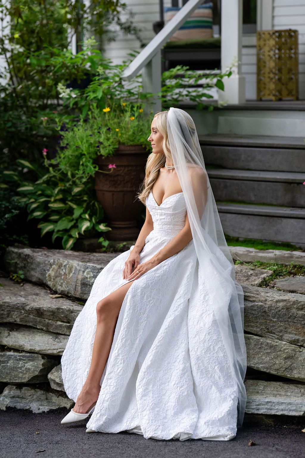 Elegant bride in white lace gown and veil sits gracefully on stone steps beside garden greenery.