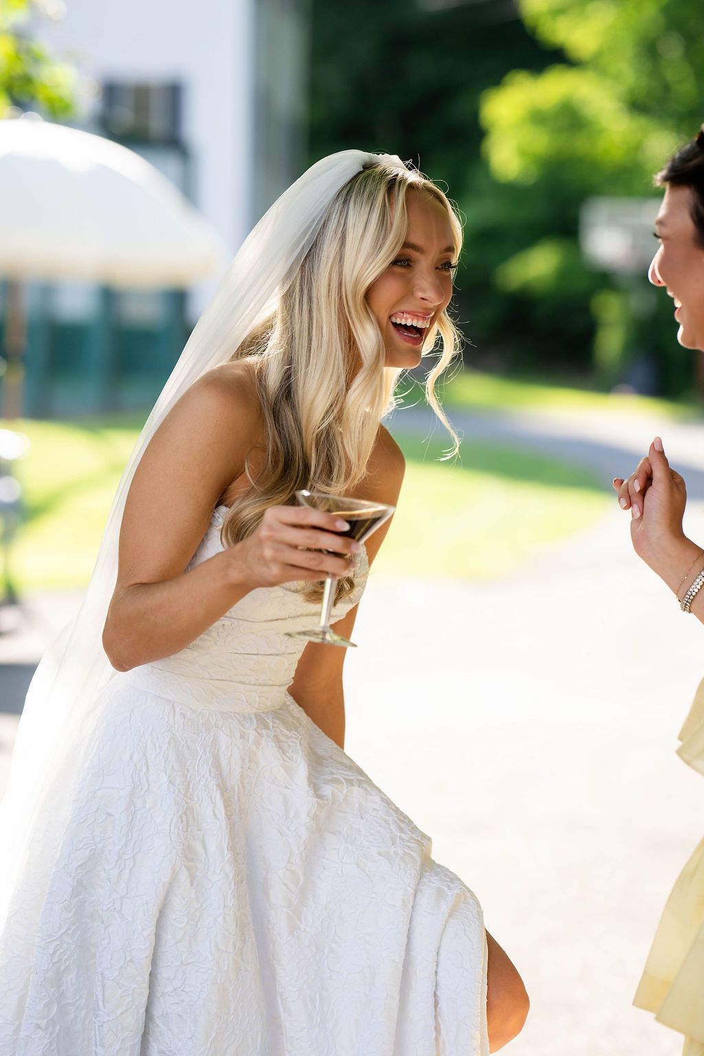 Laughing bride in strapless white gown and veil holding a cocktail glass at an outdoor wedding reception.
