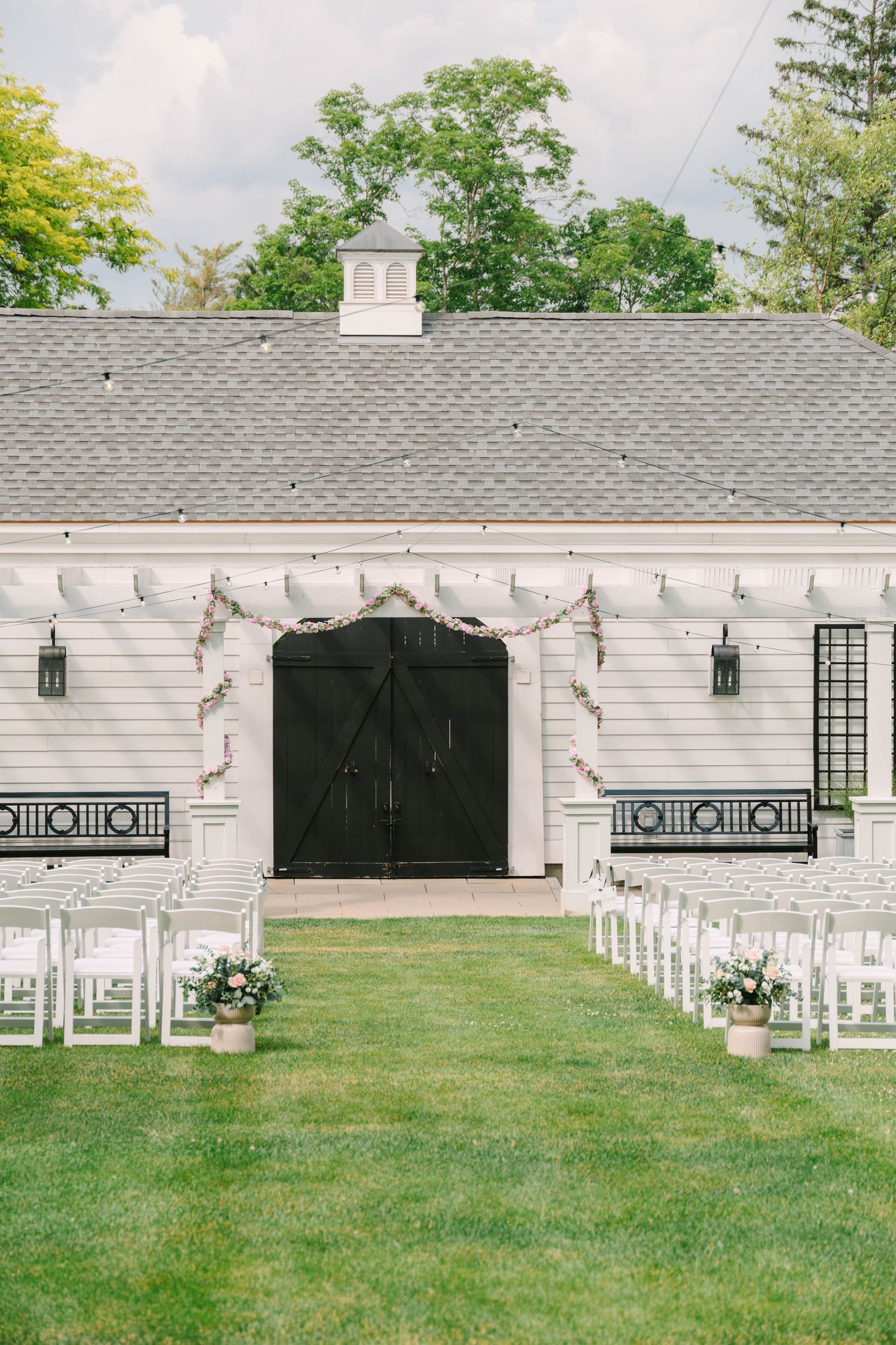 White barn wedding ceremony setup with rows of white chairs on green lawn and floral garland over black doors.