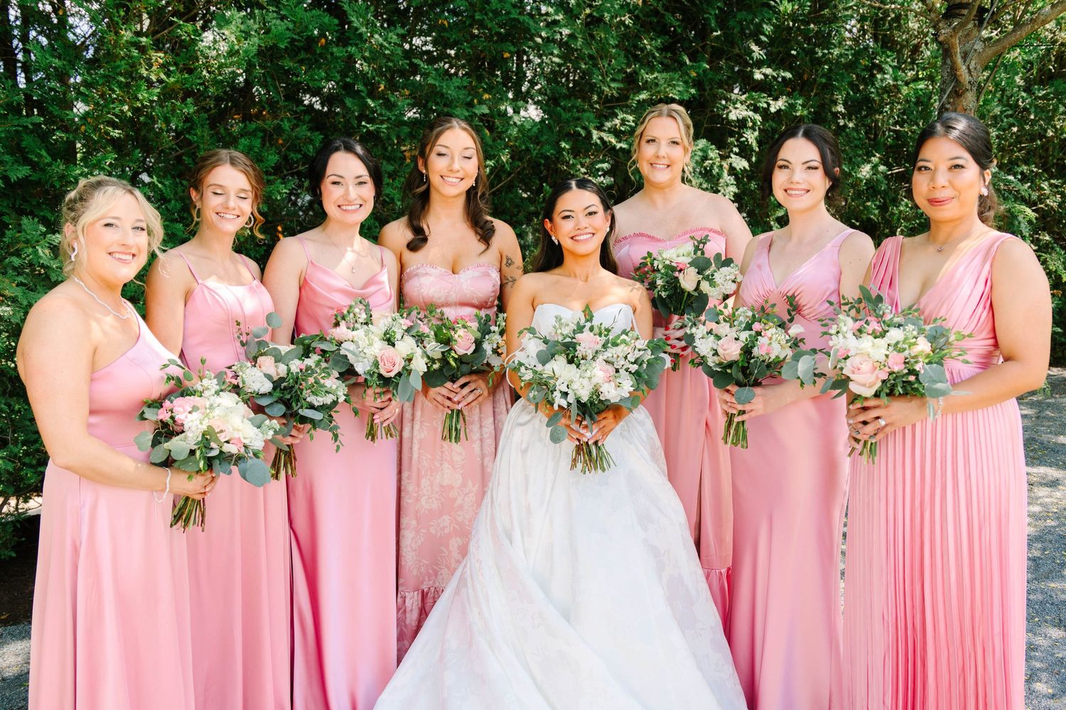 Bride in white gown surrounded by seven bridesmaids in pink dresses holding bouquets outdoors.