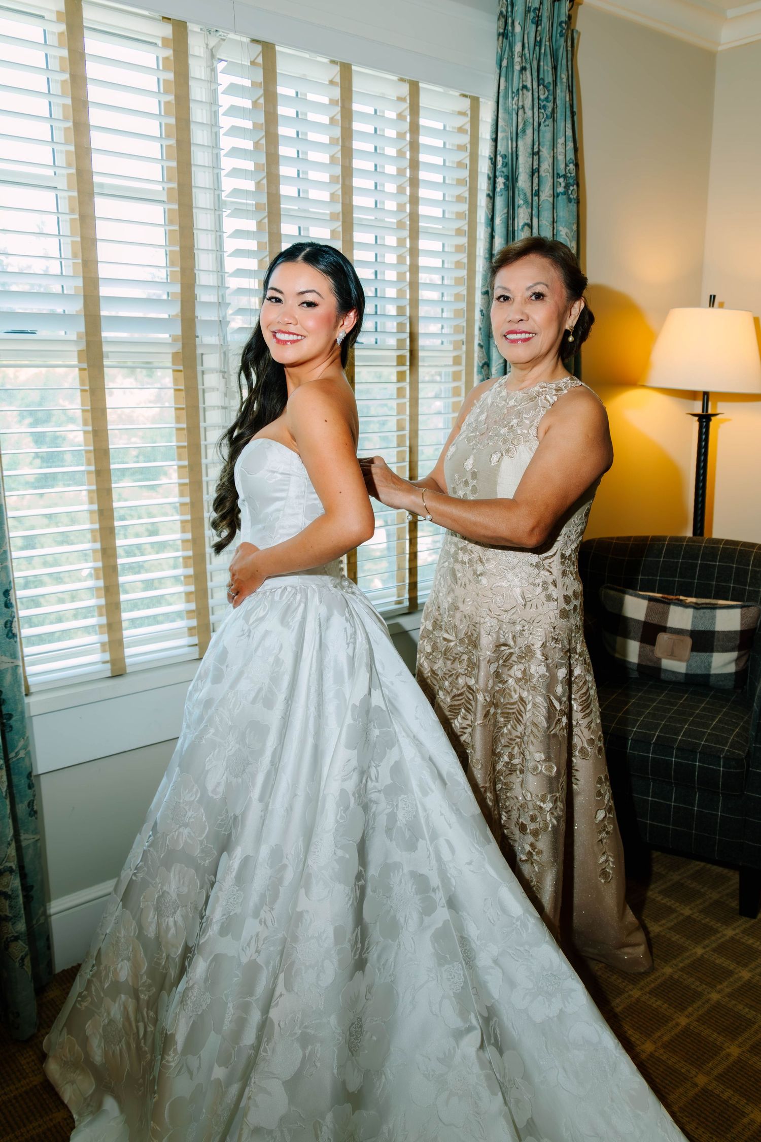 Mother helping bride into her strapless floral ballgown wedding dress near a sunlit window.